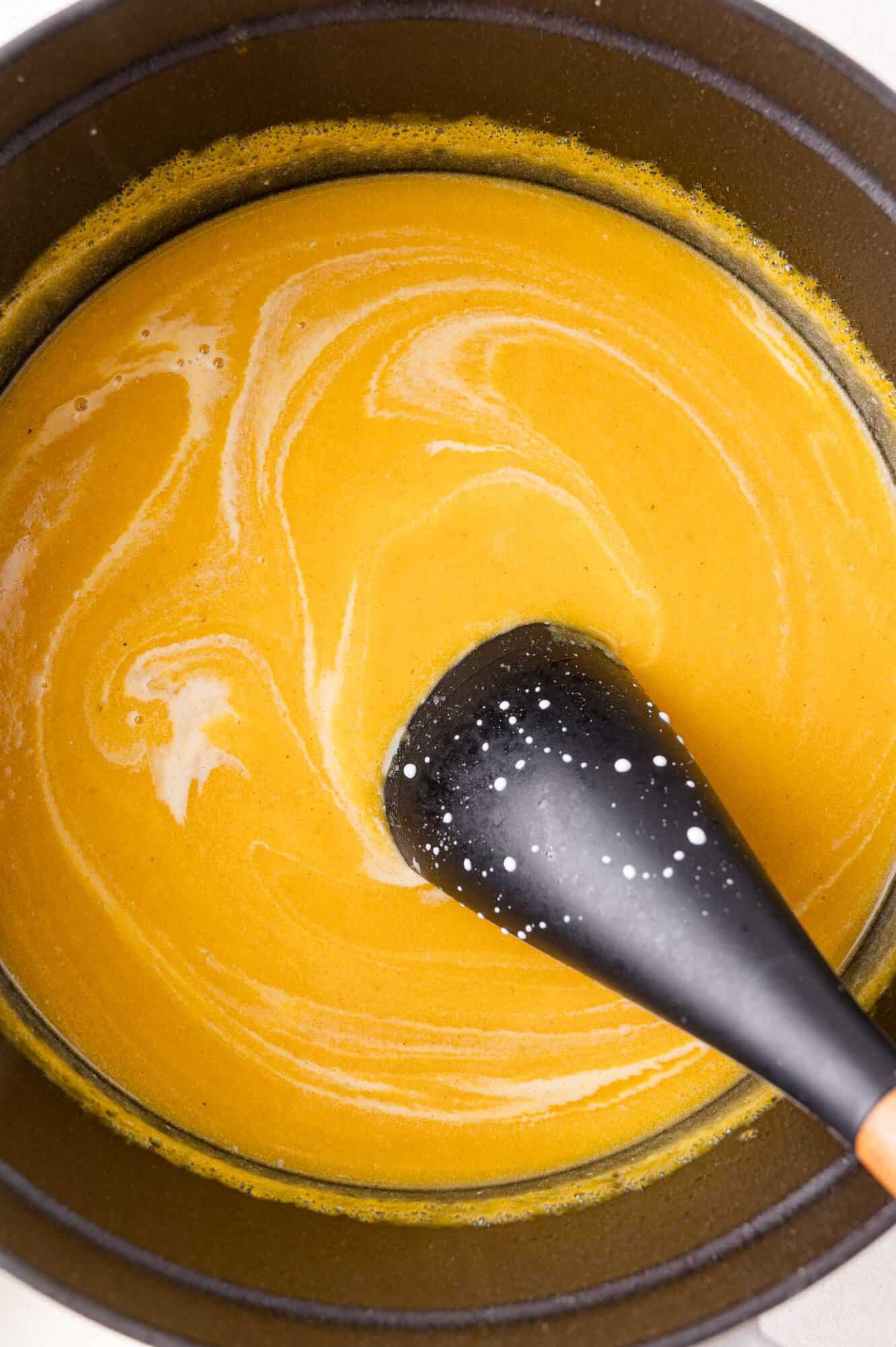 A black spoon stirs a pot of creamy squash soup, showing swirls of white liquid on the surface.