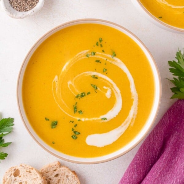 Bowl of creamy squash soup garnished with a swirl of cream and chopped herbs, next to bread slices and fresh parsley on a white surface.