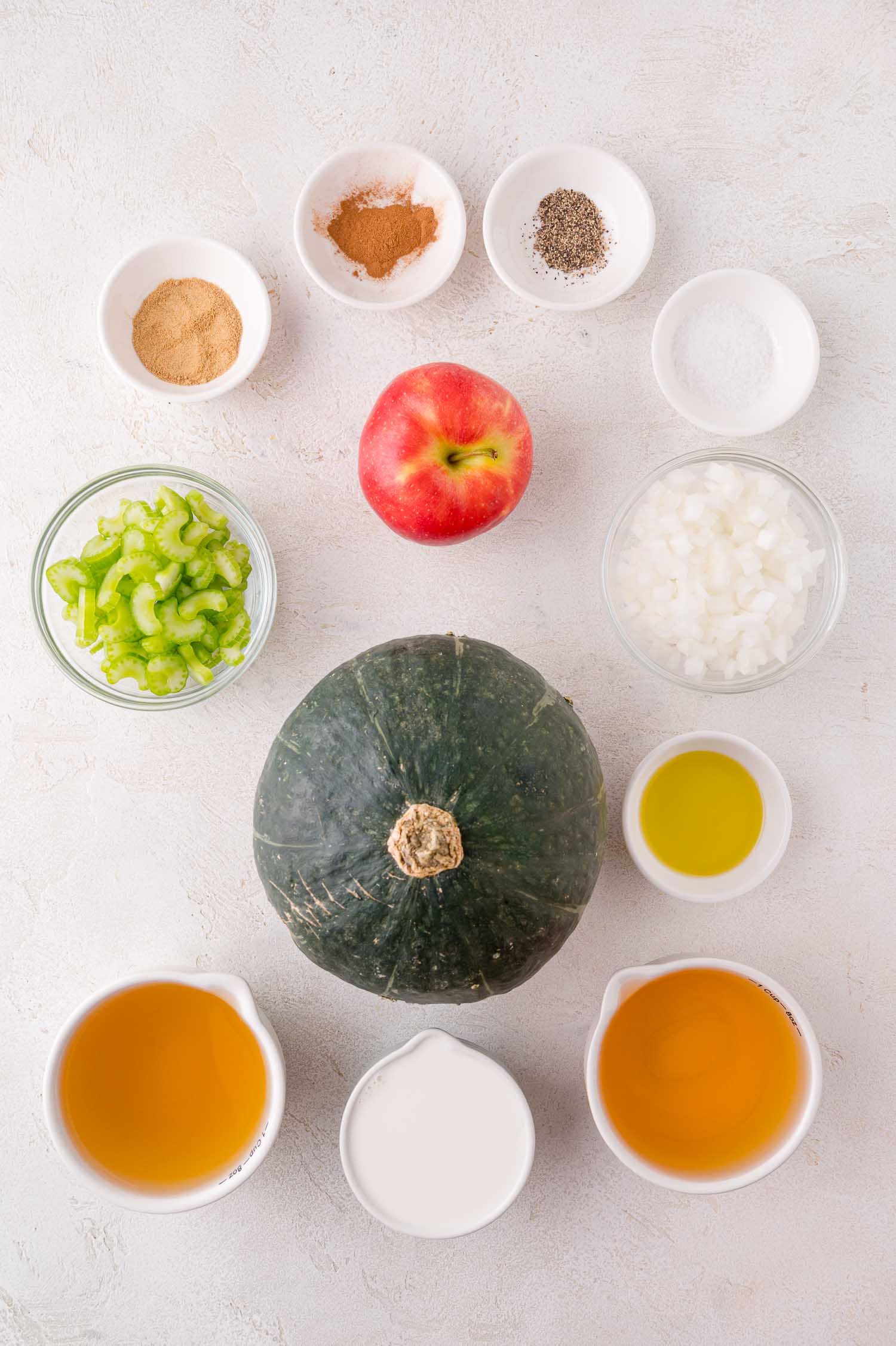 An assortment of ingredients on a white surface, including a whole kabocha squash, apple, celery, onion, spices, broth, olive oil, and cream arranged in separate bowls and cups.