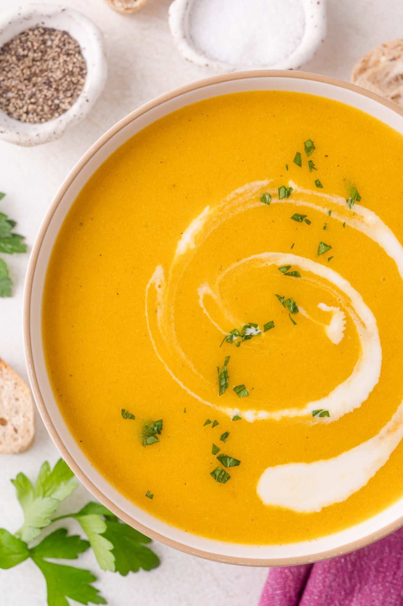 A bowl of creamy kabocha squash soup garnished with a swirl of cream and chopped herbs, surrounded by slices of bread, parsley leaves, and small dishes of salt and pepper.