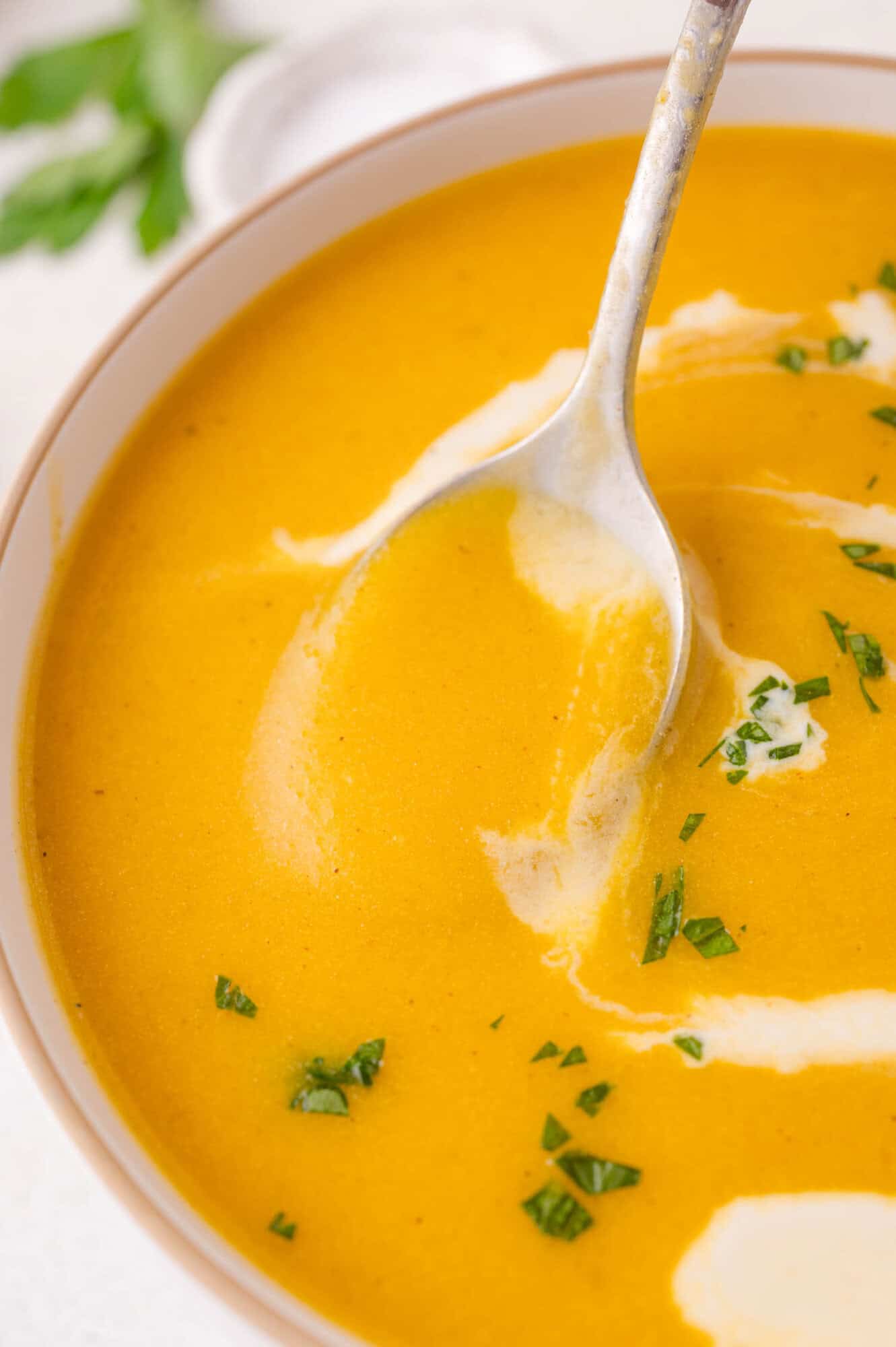 A bowl of creamy kabocha soup garnished with chopped herbs and a swirl of cream, with a spoon dipped into the soup.