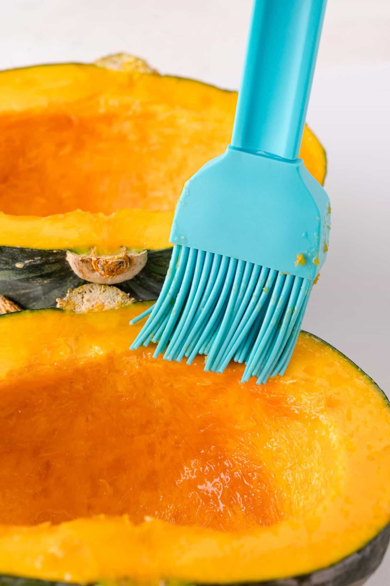 A blue silicone brush applies oil to the cut surface of a halved kabocha squash.