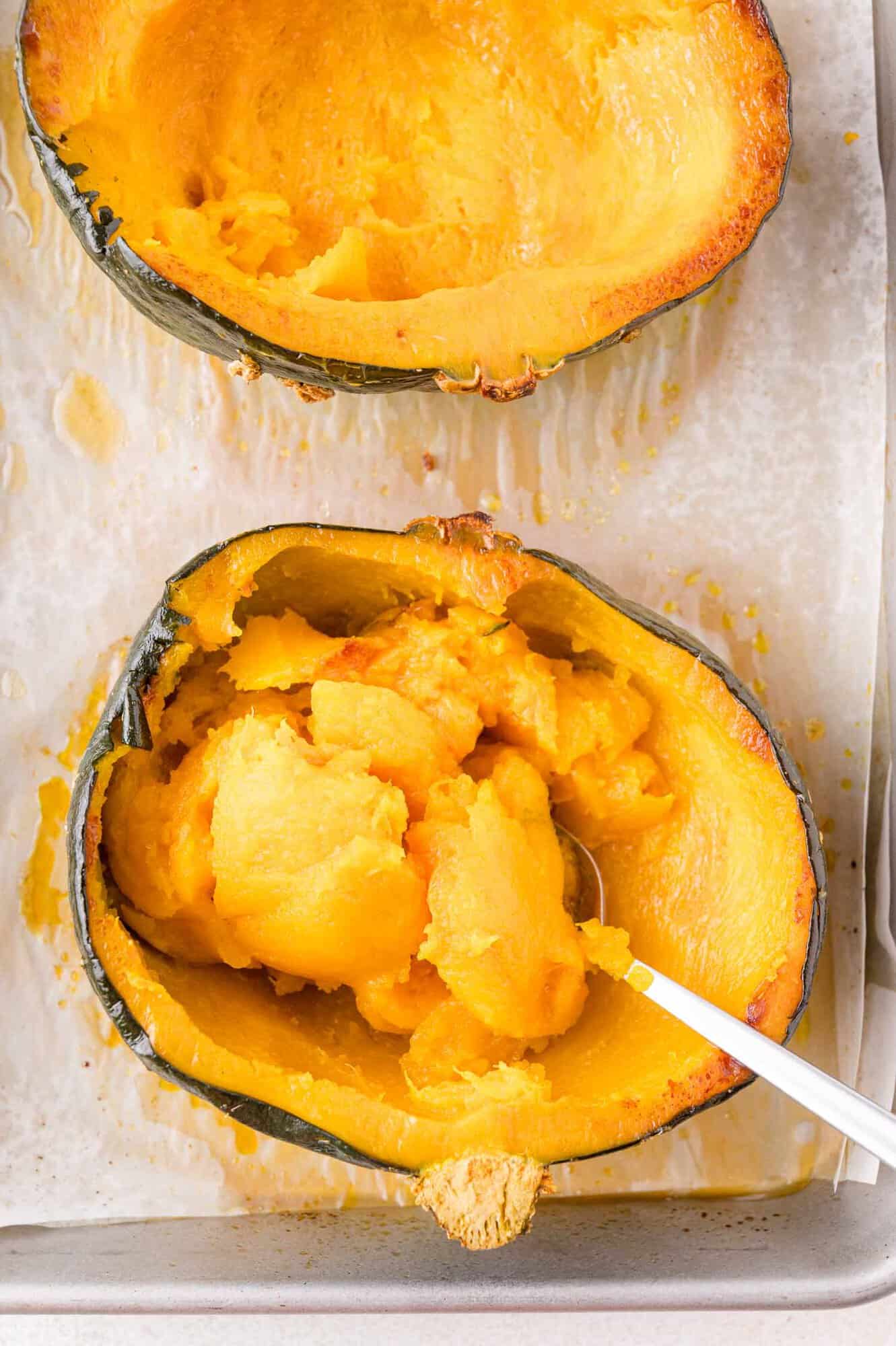 Two halves of a baked kabocha squash on a baking sheet; one half has the flesh scooped out with a spoon.