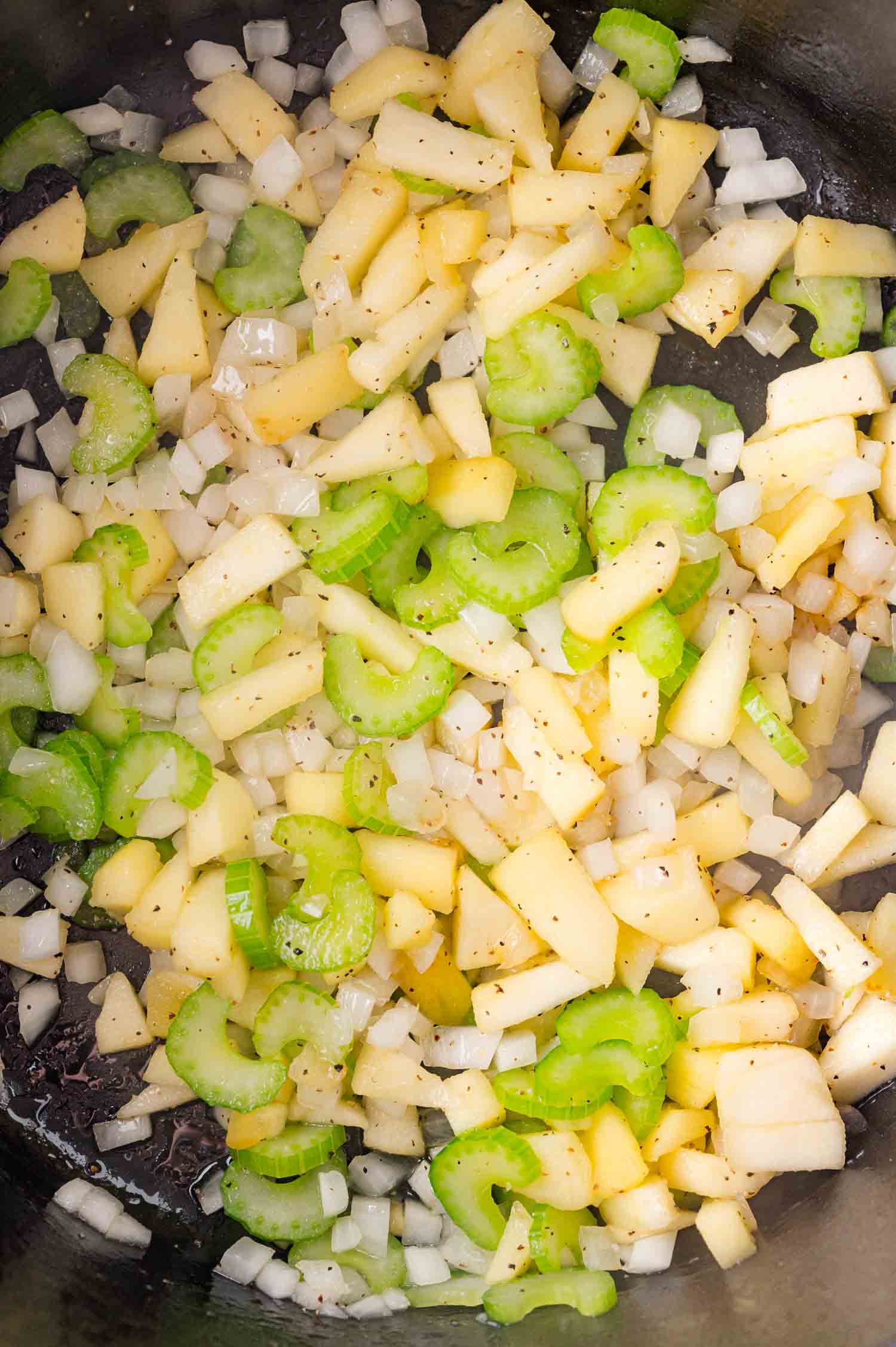 Chopped onions, celery, and apples cooking in a pan, seasoned with black pepper.