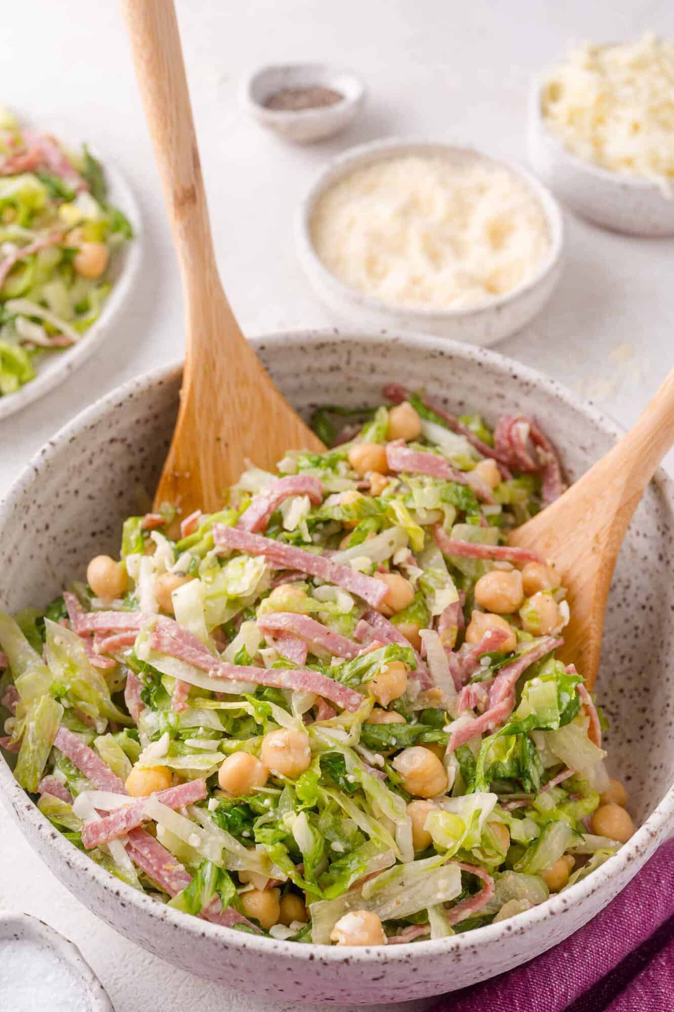 A bowl of chopped salad with lettuce, chickpeas, sliced salami, grated cheese, and two wooden salad servers. Small dishes with cheese and pepper are in the background.