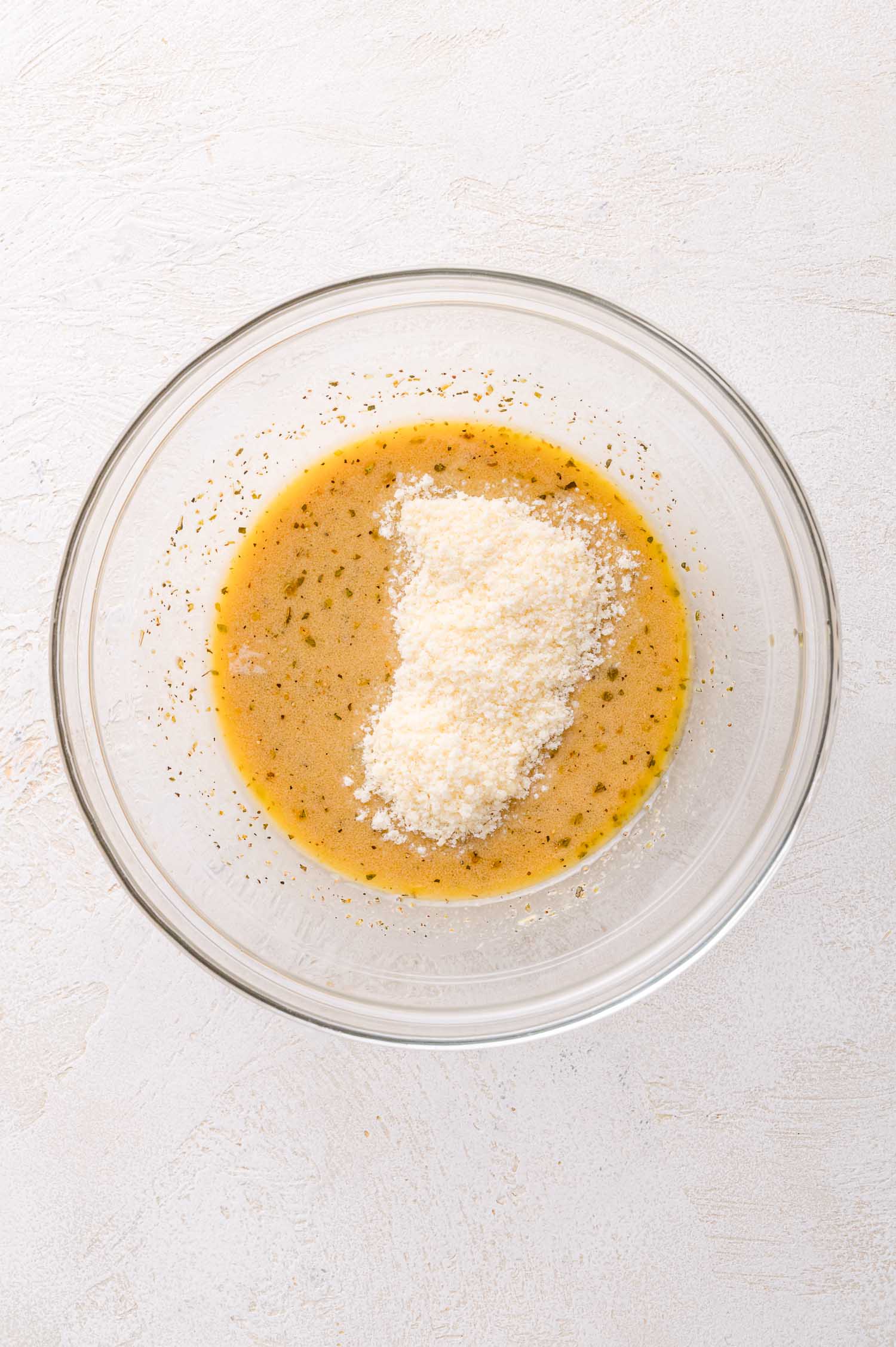 A glass bowl containing a dressing mixture with a mound of grated parmesan cheese on top, placed on a light-colored surface.