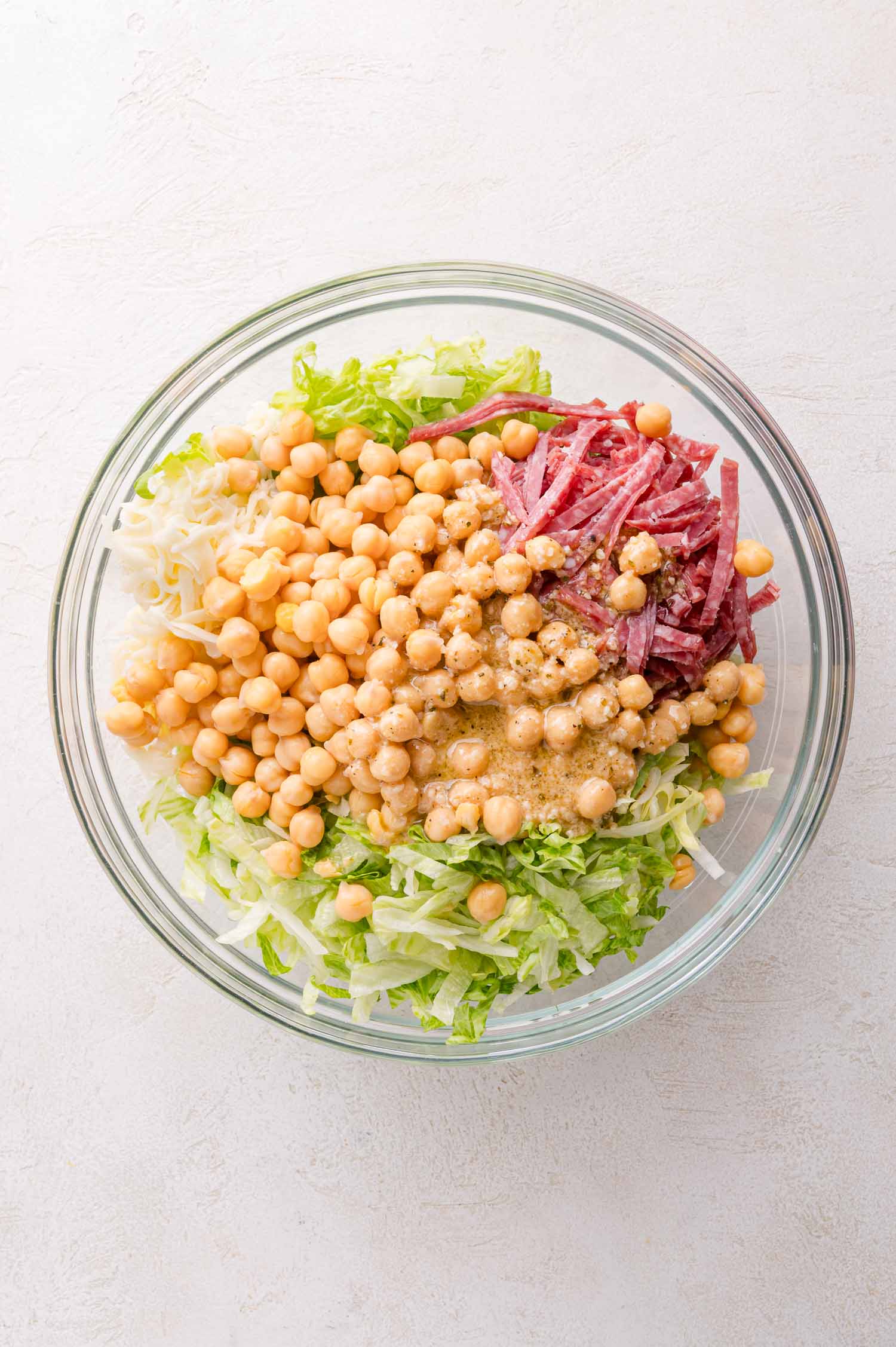 A glass bowl filled with shredded lettuce, chickpeas, shredded cheese, sliced salami, and dressing on a light background.