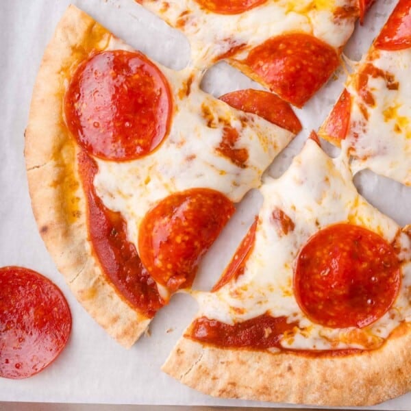 A pepperoni pita pizza with melted cheese, sliced into pieces, on a sheet of parchment paper with one pepperoni slice off to the side.