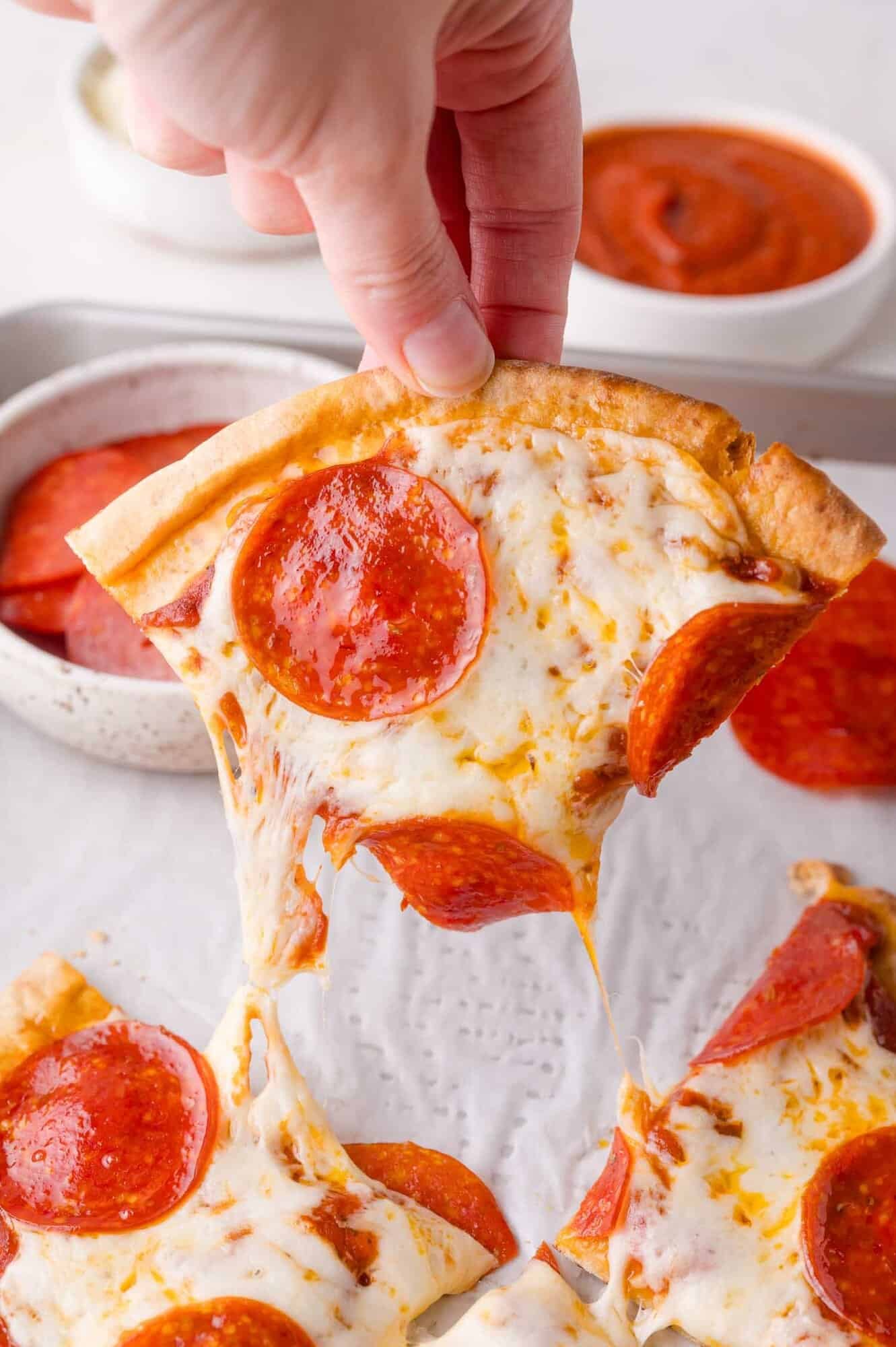 A hand lifts a slice of pepperoni pizza with melted cheese stretching, with more pizza and bowls of pepperoni and sauce in the background.