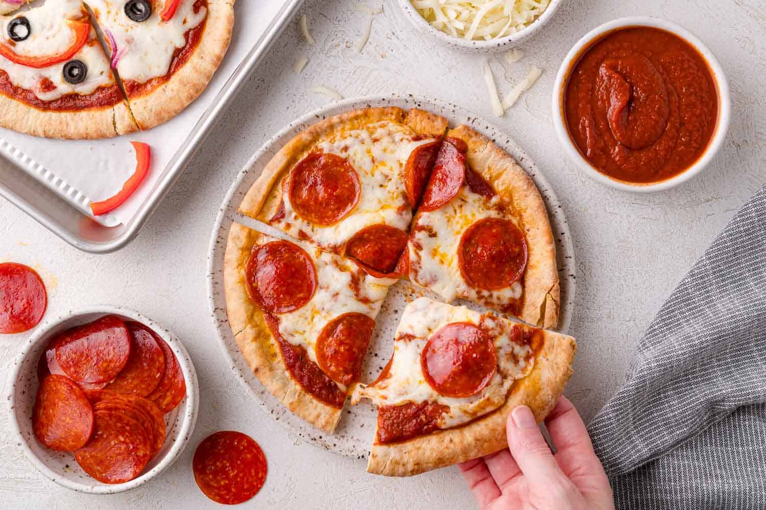 A hand takes a slice of pepperoni pita pizza from a plate, surrounded by a tray of veggie pizza, pepperoni slices, shredded cheese, and a bowl of pizza sauce.