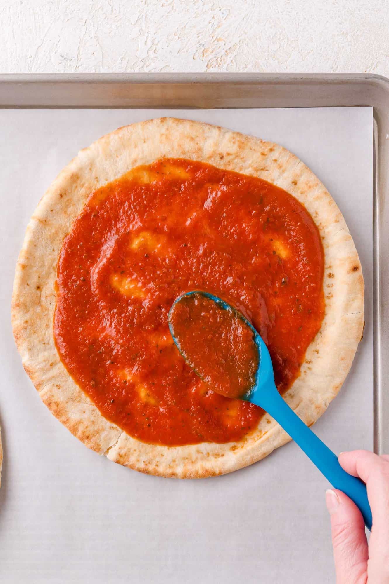 A hand spreads tomato sauce on a round pita with a blue spoon, on a parchment-lined baking sheet.