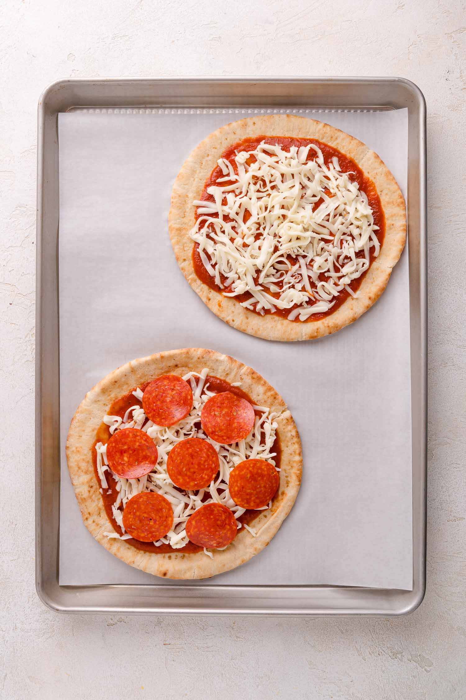 Two round, uncooked pita pizzas on a baking sheet; one topped with cheese only, the other with cheese and pepperoni slices.