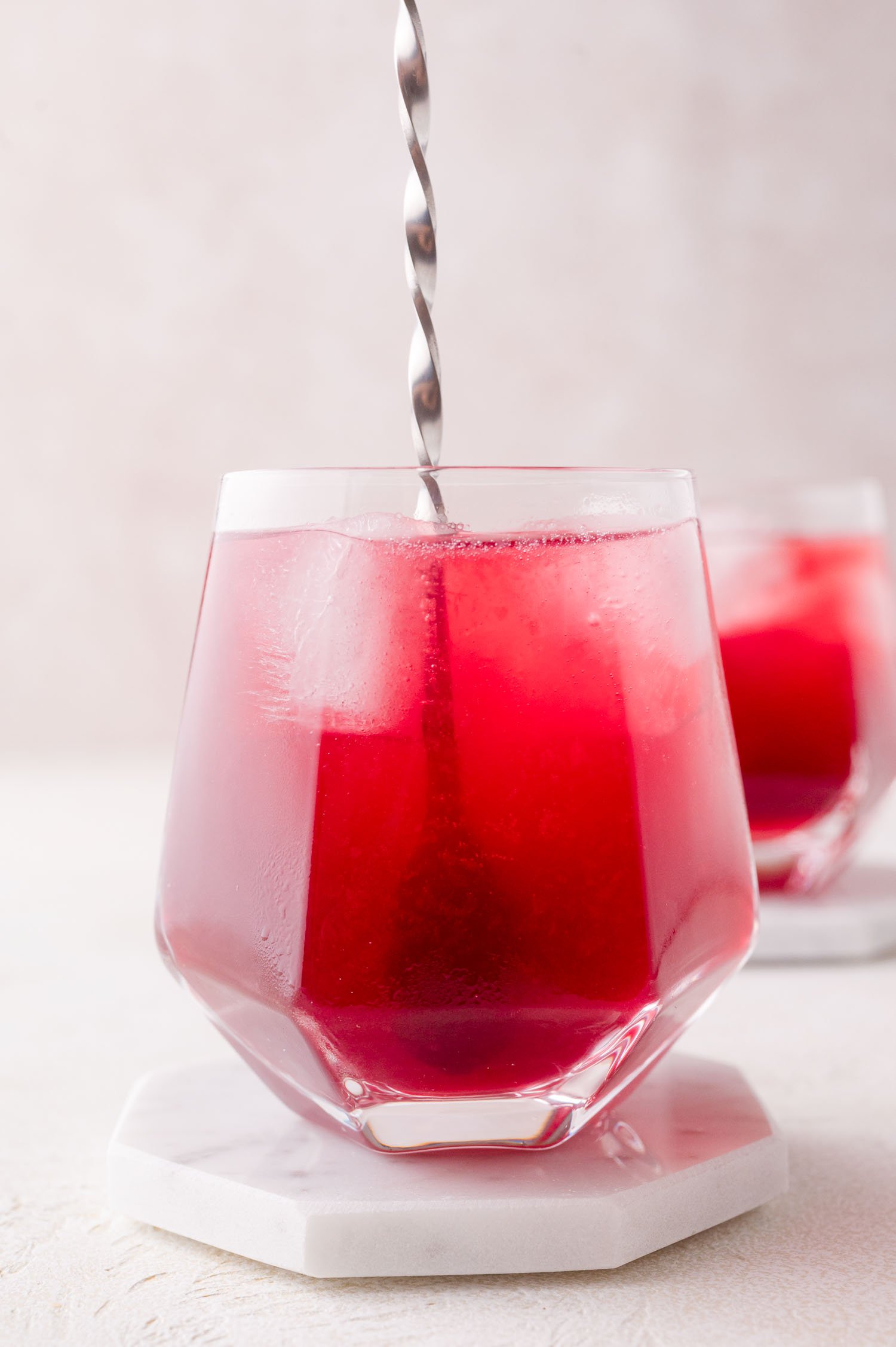 A glass of pomegranate spritzer being stirred with a metal spoon, placed on a white hexagonal coaster, with another similar glass blurred in the background.