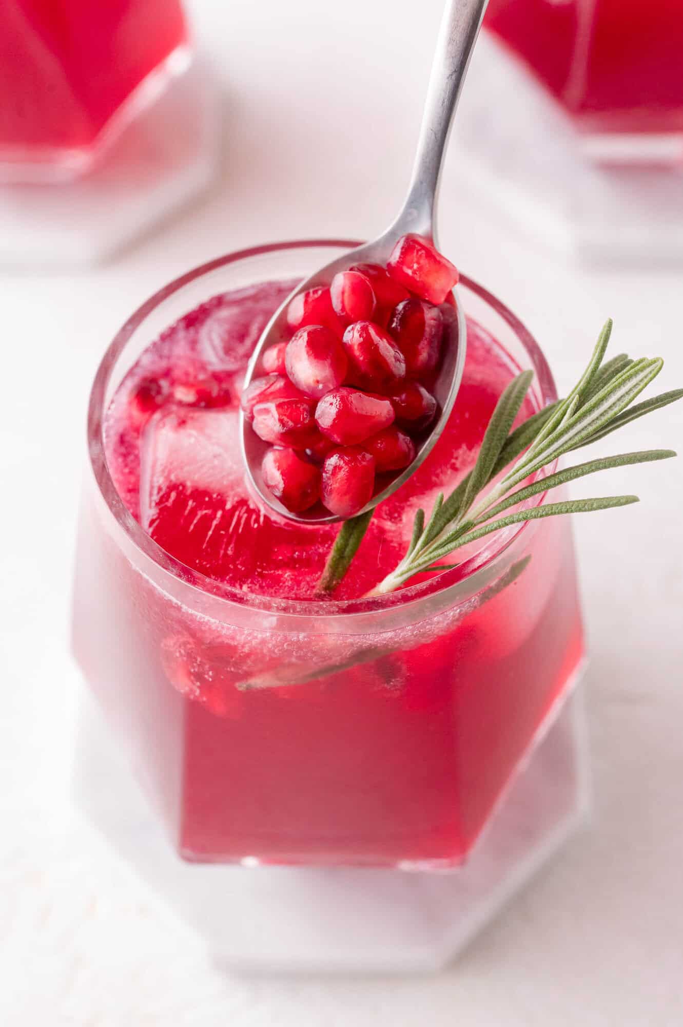 A glass of red pomegranate drink with ice, garnished with a rosemary sprig and pomegranate seeds on a spoon above the glass.