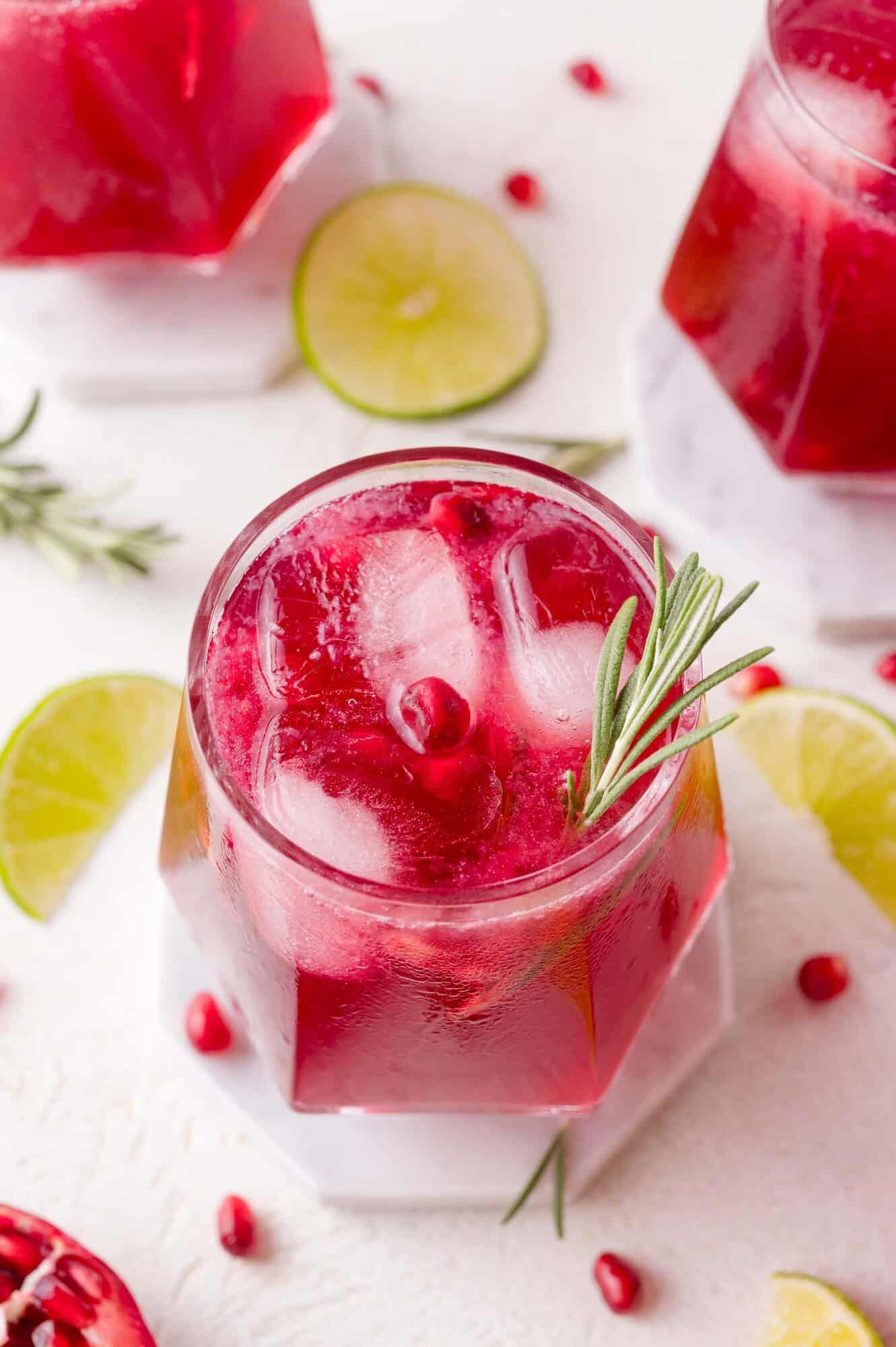 A glass of red pomegranate mocktail with ice, garnished with a sprig of rosemary, surrounded by lime slices and pomegranate seeds on a white surface.