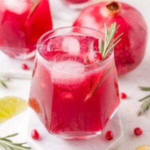 A glass of red pomegranate spritzer with ice and a rosemary sprig garnish sits on a coaster, with a whole pomegranate and lime slices in the background.