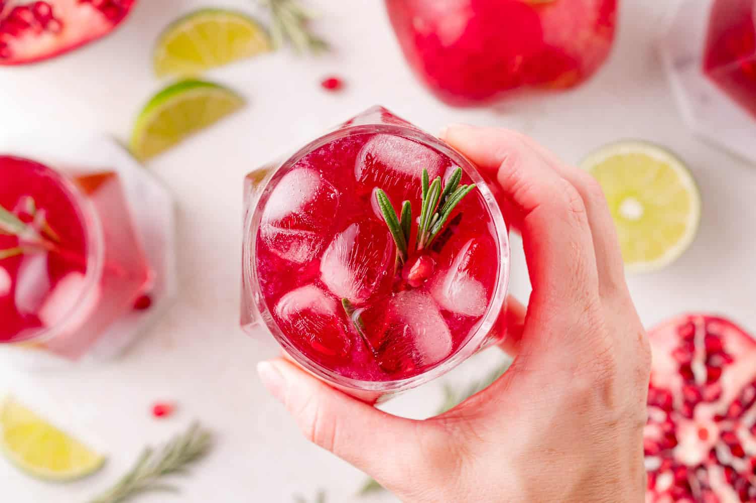 A hand holds a glass of pomegranate spritzer garnished with rosemary and pomegranate seeds; lime slices and pomegranate pieces are visible on the table.