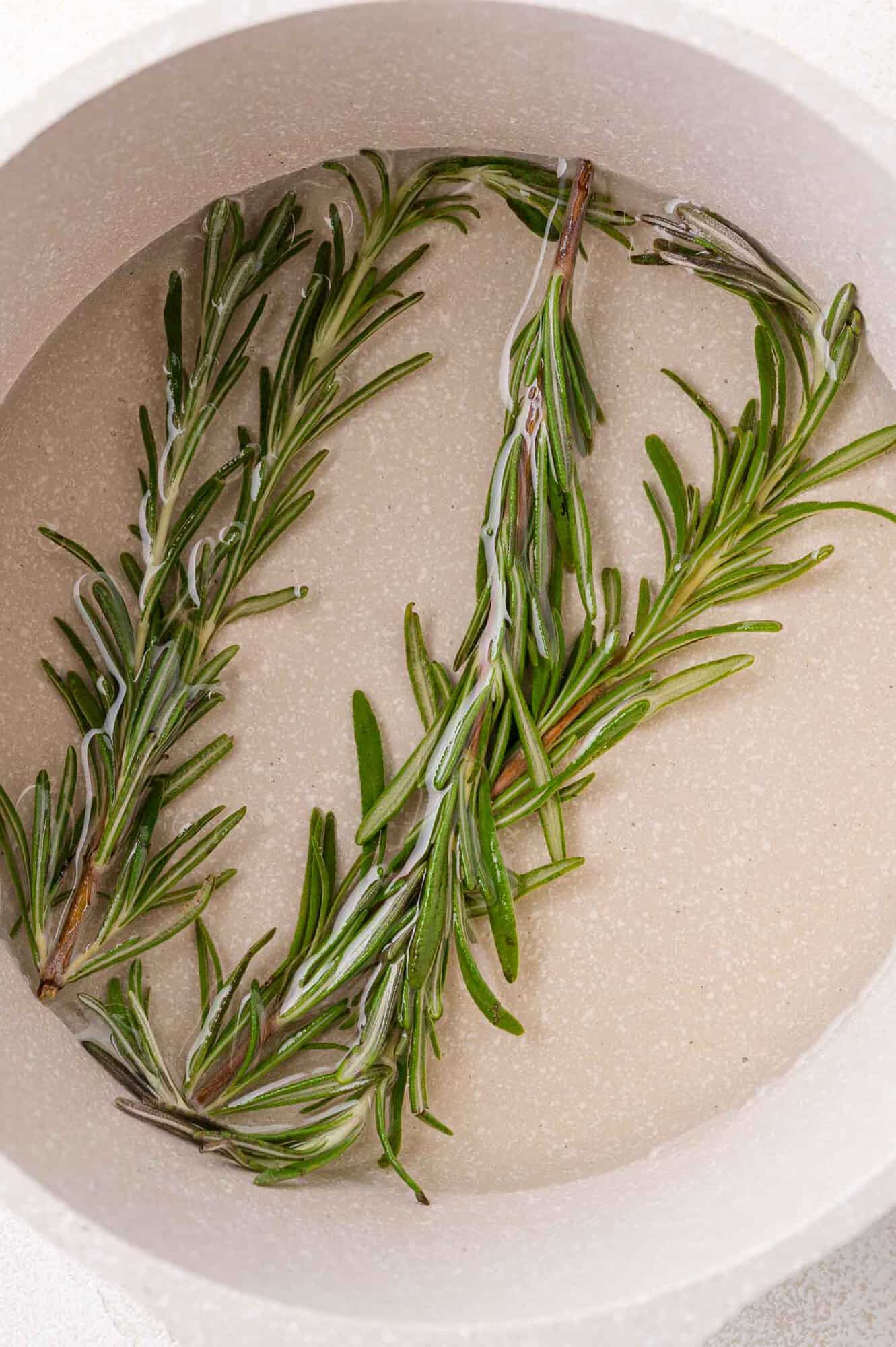 Two sprigs of fresh rosemary submerged in simple syrup inside a light-colored pot.