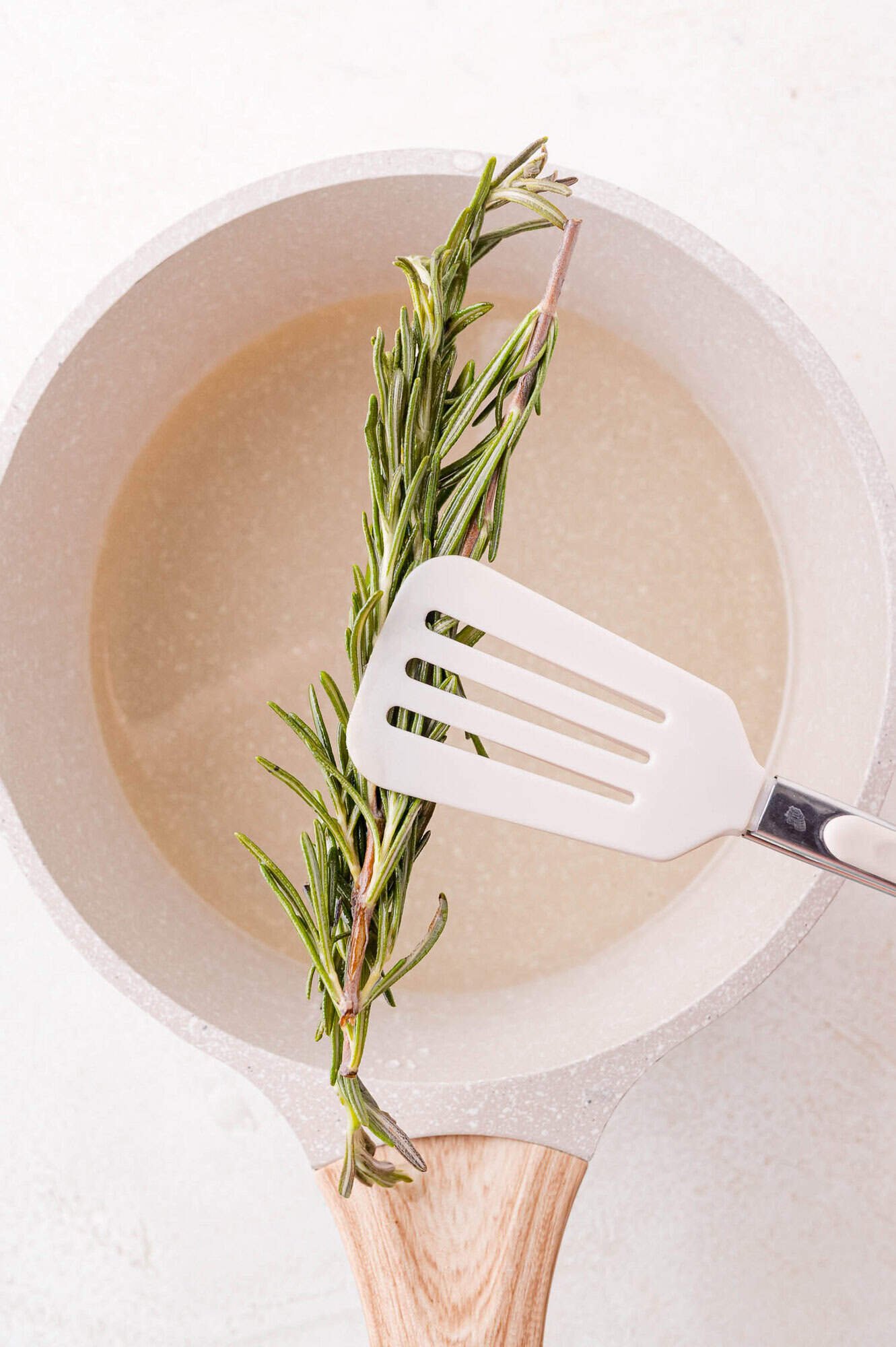 A sprig of fresh rosemary above a saucepan with simple syrup, being lifted up with a white spatula.