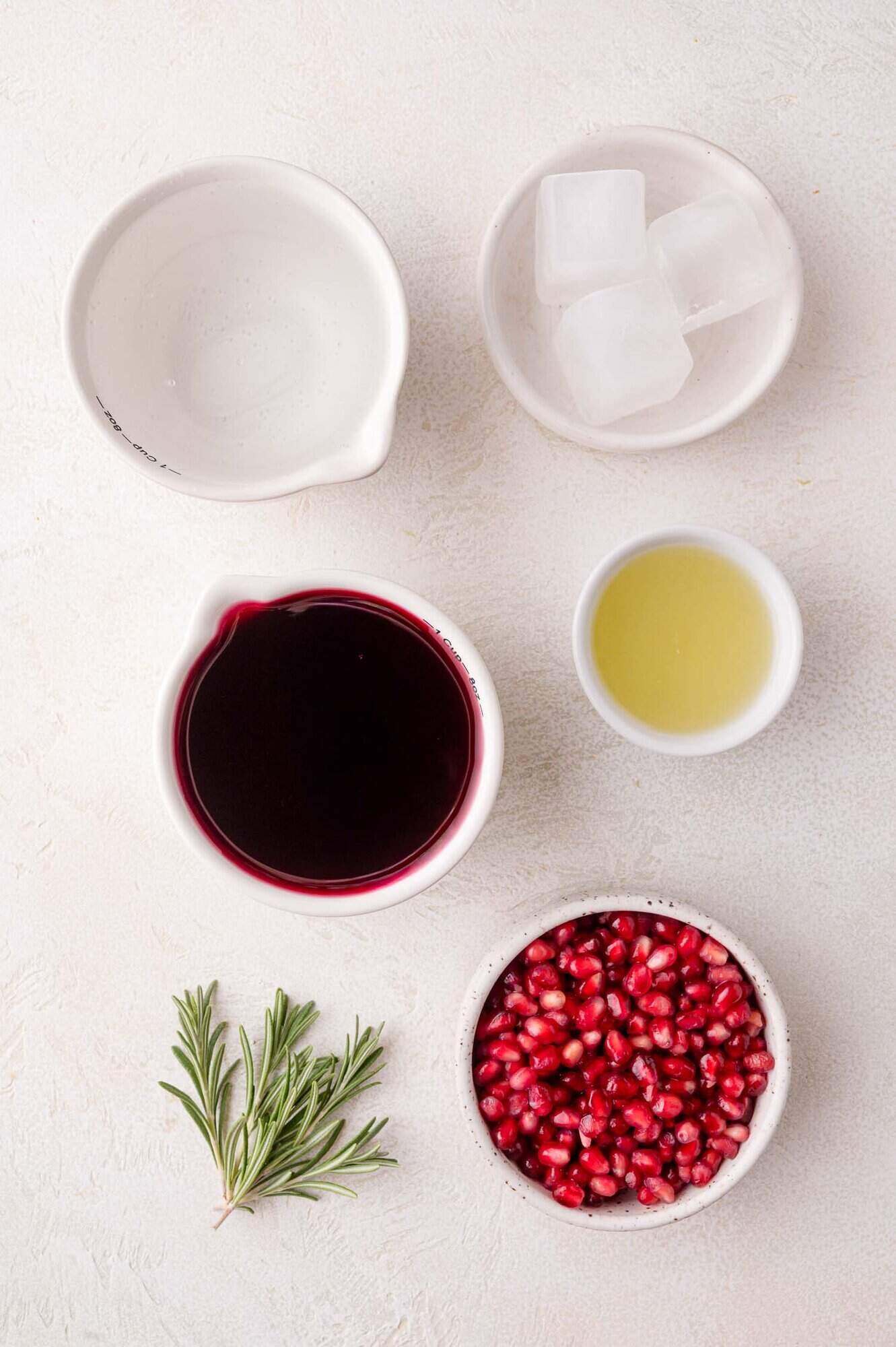 Five bowls hold pomegranate juice, pomegranate seeds, lime juice, ice cubes, and a sprig of rosemary arranged on a white surface.