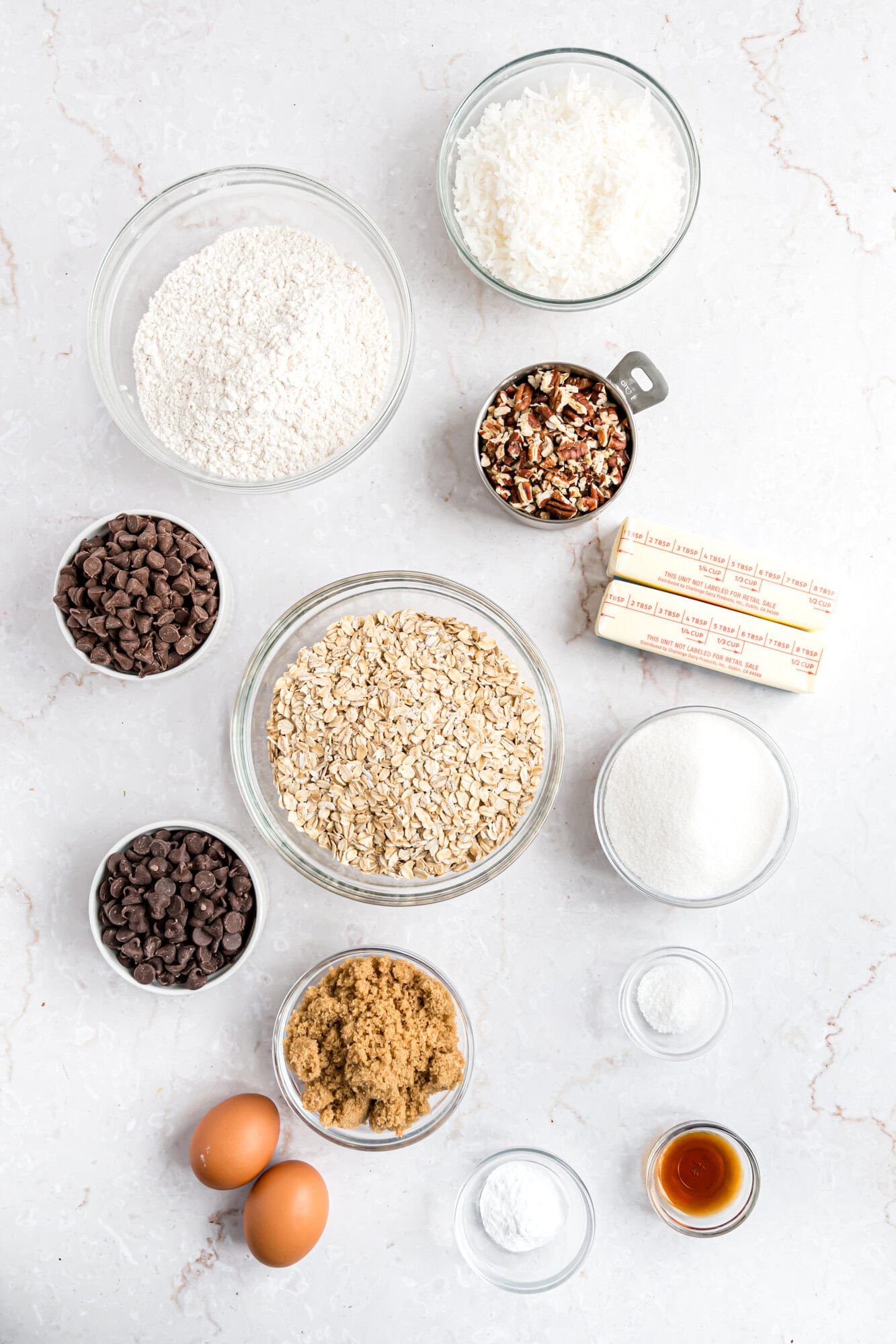 Various baking ingredients in clear bowls on a white surface, including flour, oats, shredded coconut, pecans, chocolate chips, sugar, brown sugar, eggs, baking soda, vanilla, and butter.
