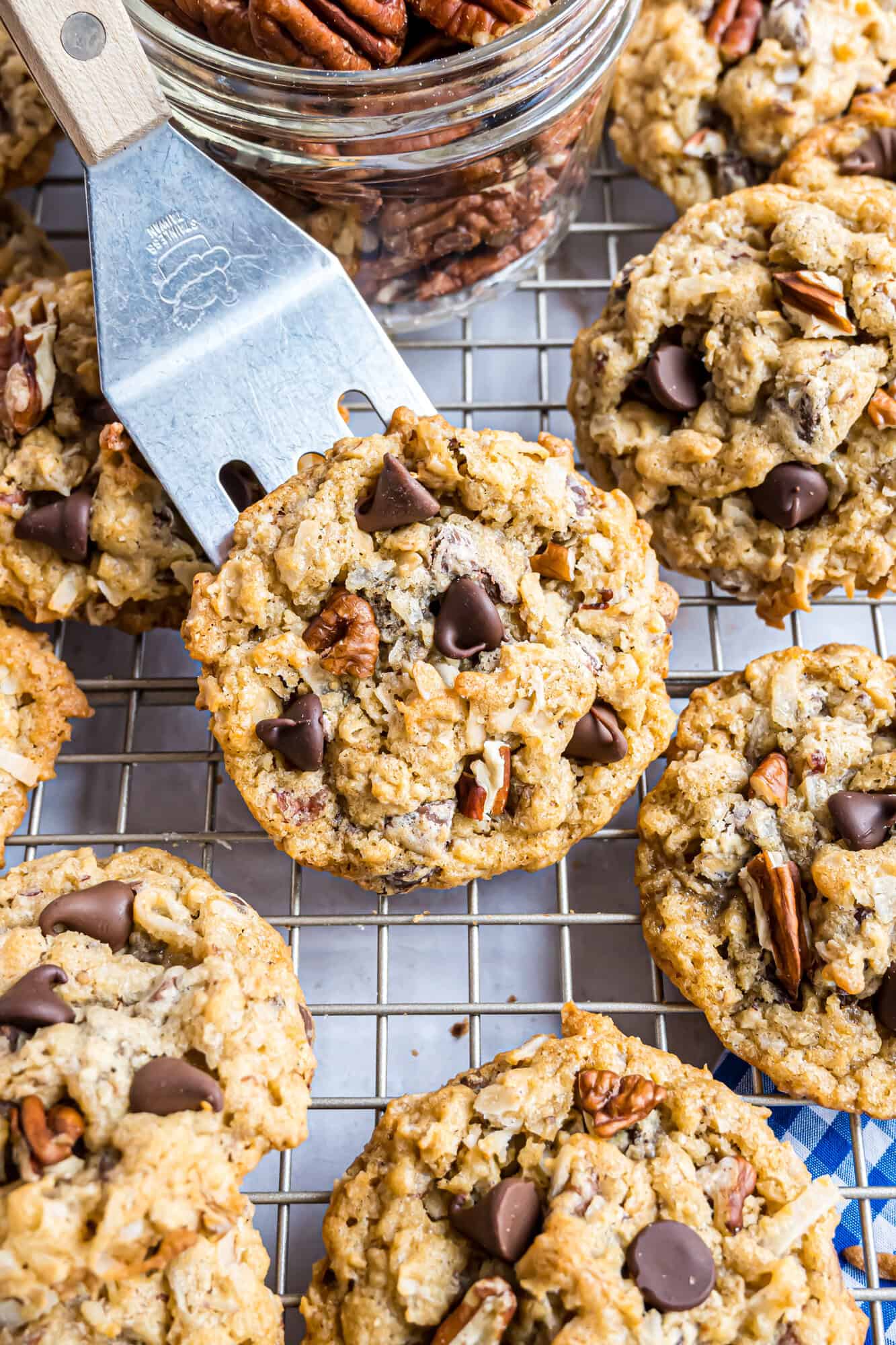Cowboy cookies with chocolate chips and pecans on a cooling rack, with a spatula lifting one and a jar of pecans nearby.