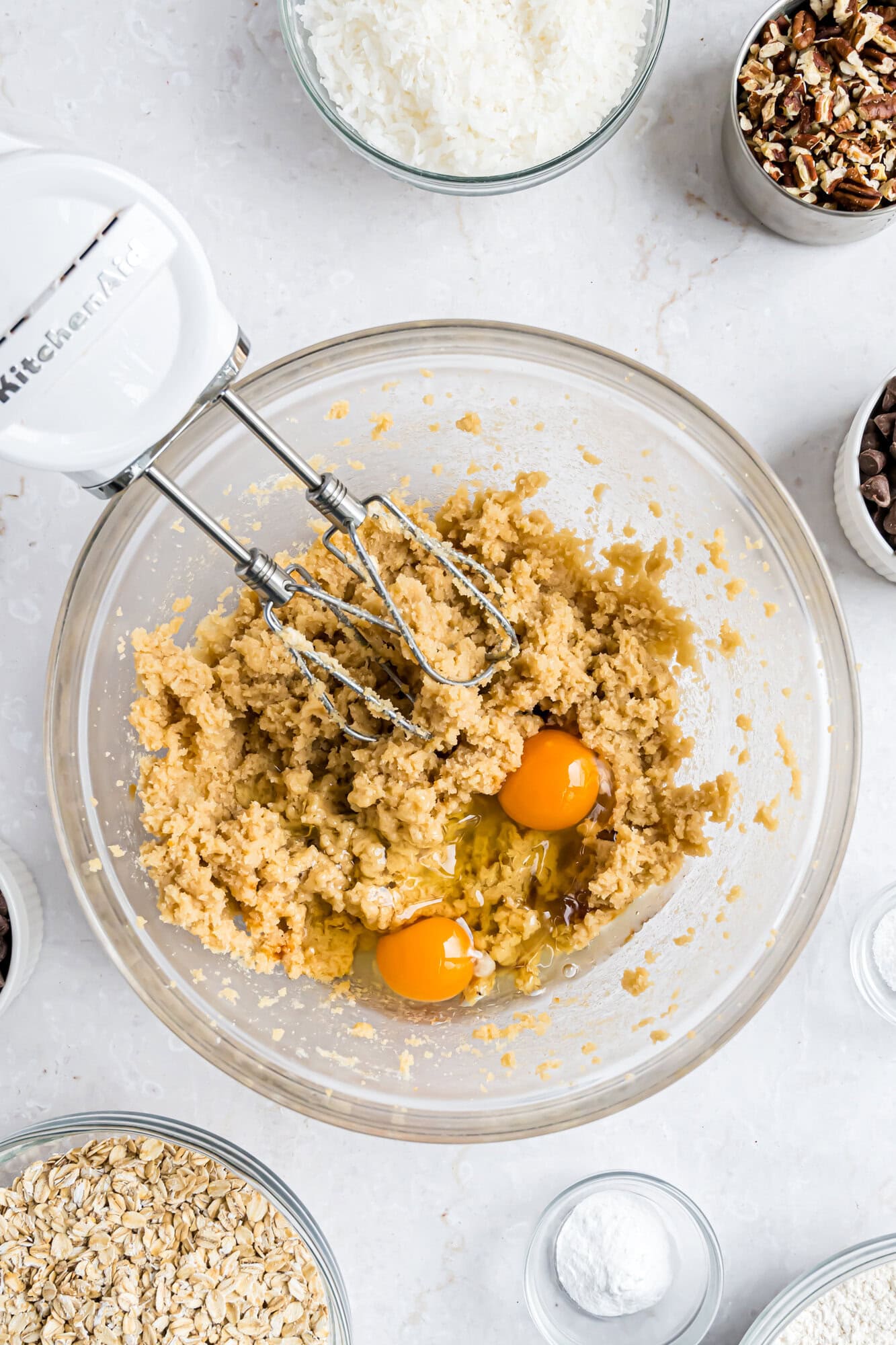 A glass bowl with cookie dough, two eggs, and a hand mixer surrounded by small bowls of oats, coconut, chocolate chips, pecans, and baking powder on a white surface.