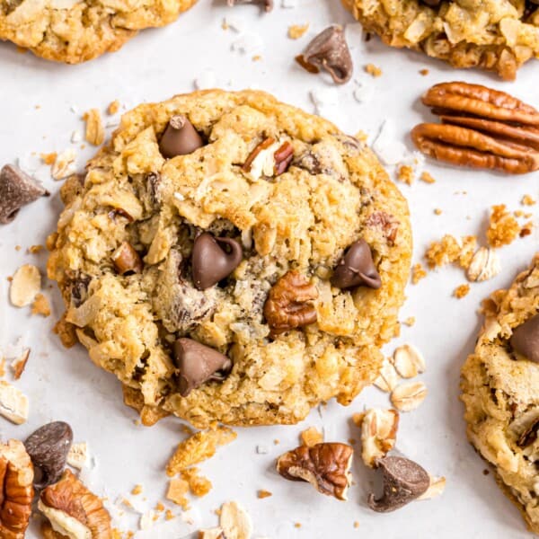 Cowboy cookie with chocolate chips and pecans on a white surface, surrounded by scattered oats, pecans, and chocolate chips.