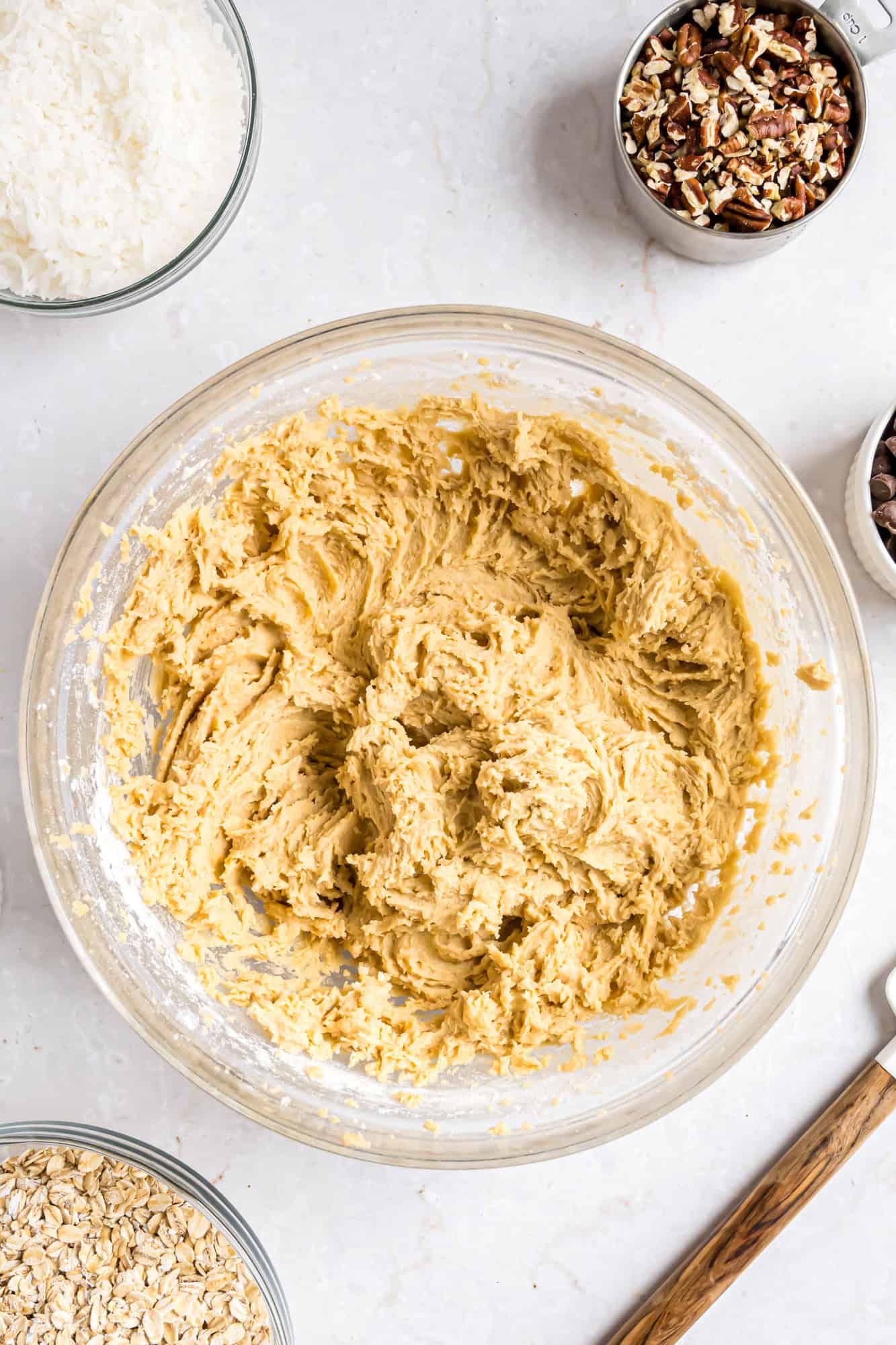 A glass bowl filled with cookie dough sits on a countertop, surrounded by bowls of chopped pecans, oats, shredded coconut, and chocolate chips.
