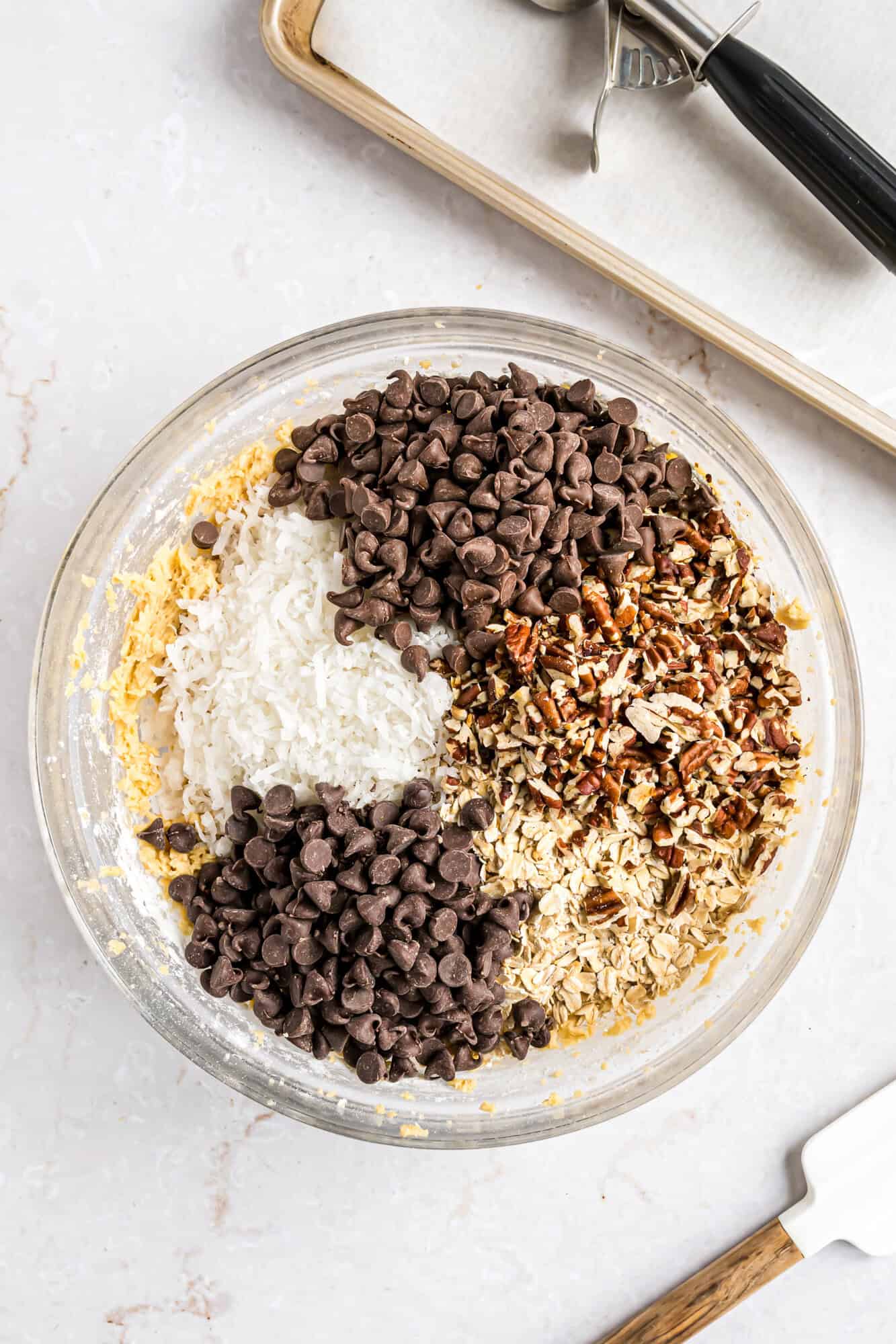 A glass bowl filled with chocolate chips, shredded coconut, chopped nuts, and oats, ready to be mixed; a baking tray and spatula are nearby.