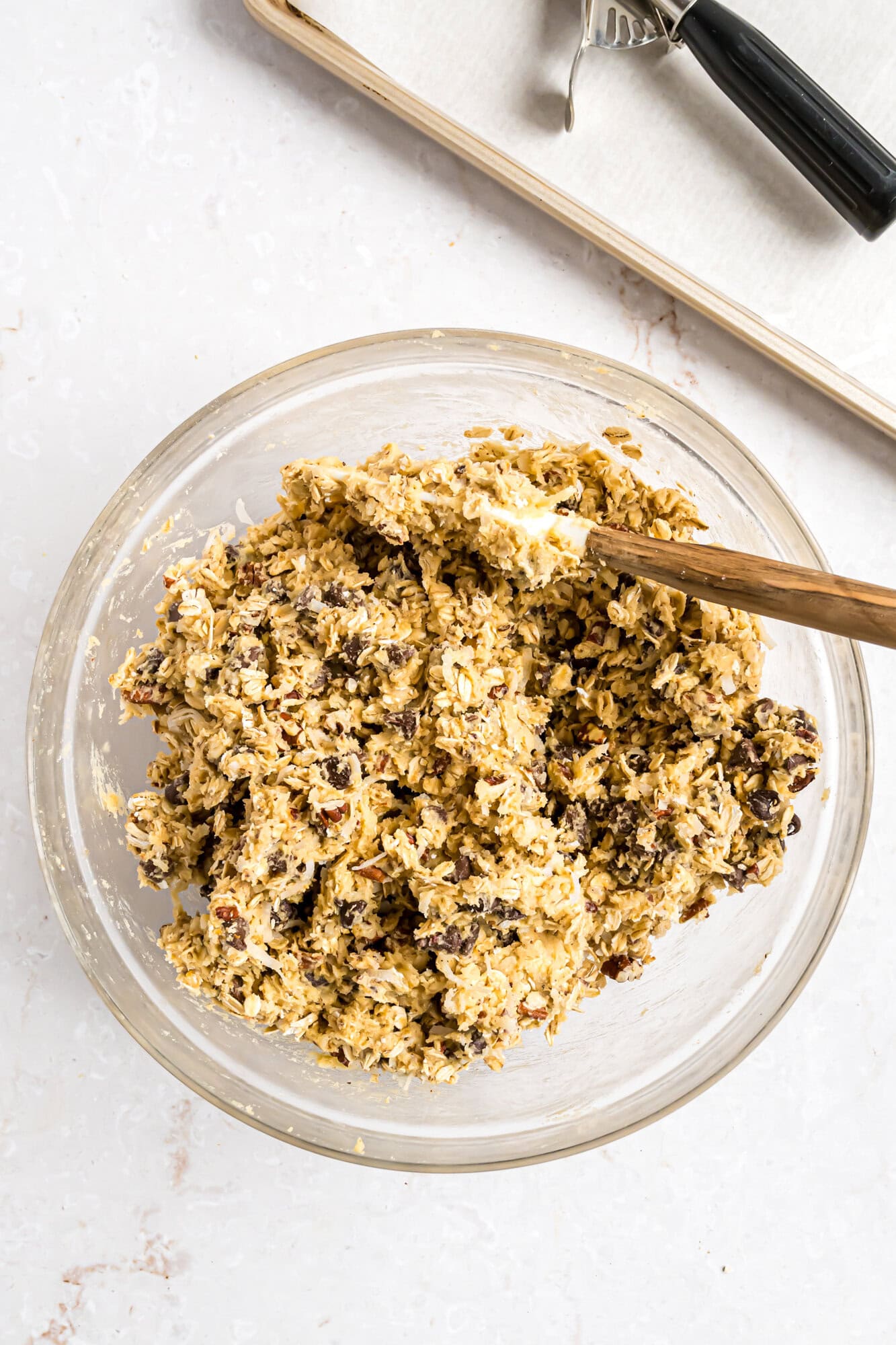 A glass bowl filled with cookie dough mixed with chocolate chips and nuts, with a wooden spoon resting inside. A baking sheet and cookie scoop are in the background.