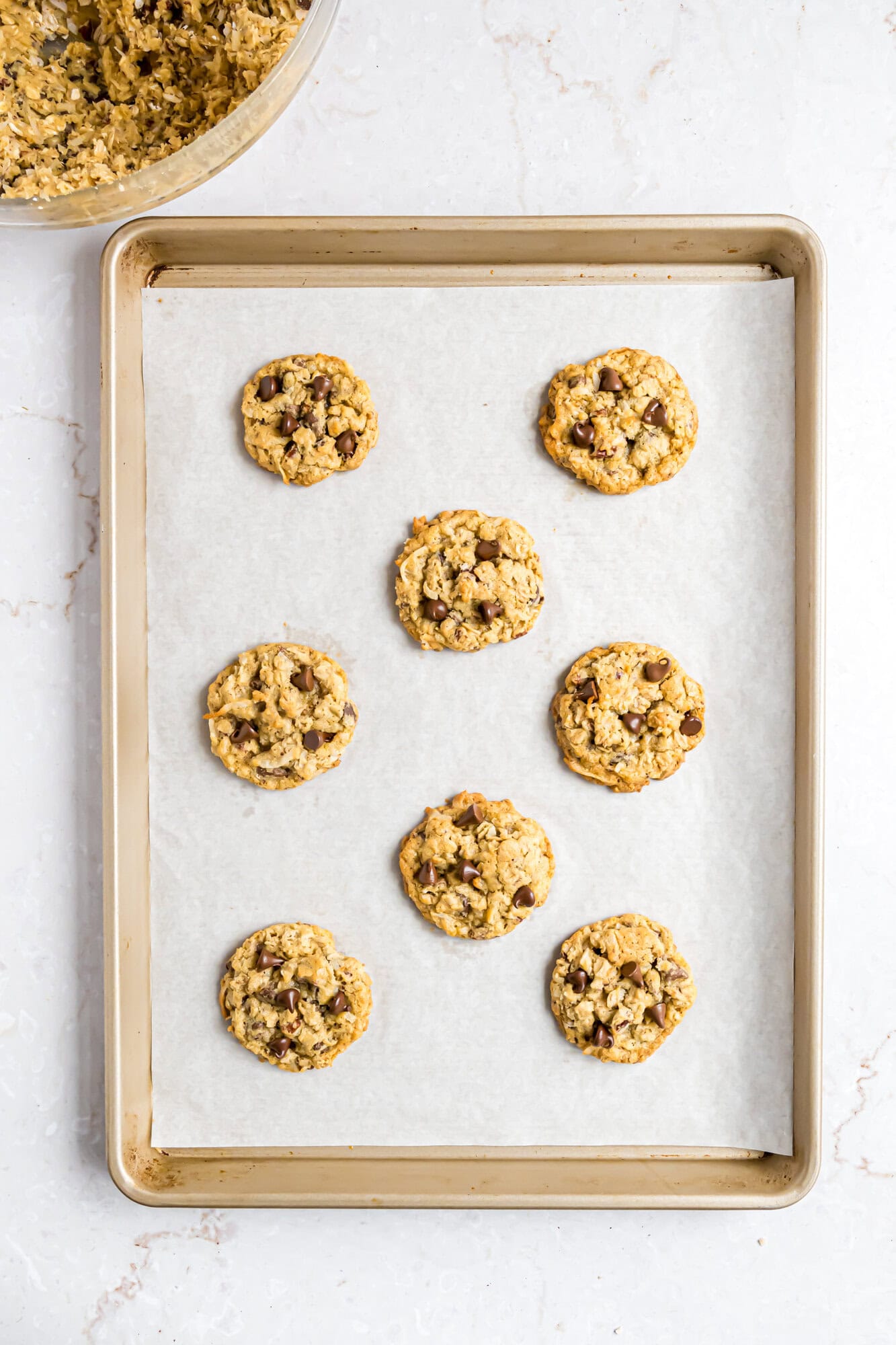 A baking sheet lined with parchment paper holds eight baked cowboy cookies, with more cookie dough visible in a mixing bowl at the top left corner.