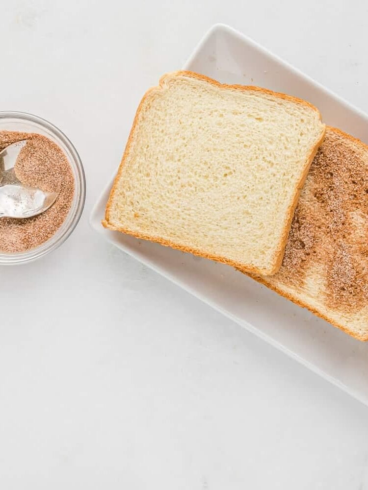 Bread slices on a plate dusted with cinnamon and sugar, next to a small bowl of cinnamon sugar with a spoon.