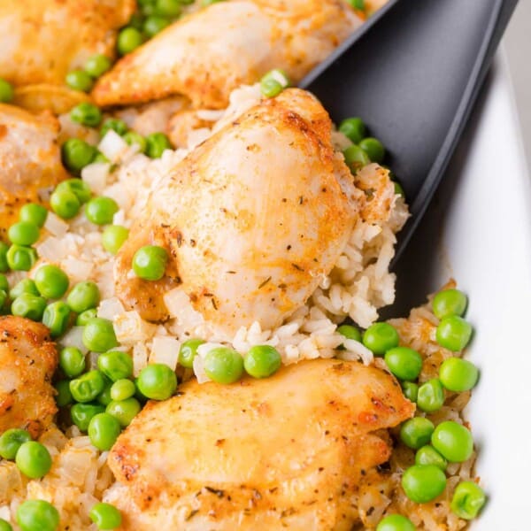 A serving spoon scooping seasoned chicken pieces, rice, and green peas from a white dish.