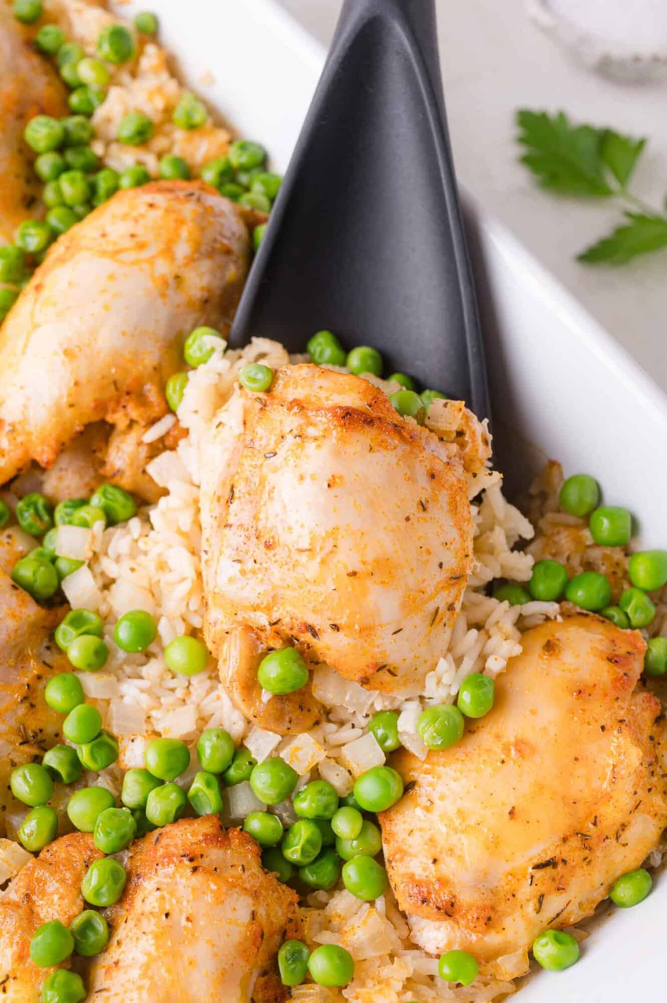 A serving spoon lifts a portion of baked chicken thighs, rice, and green peas from a white dish.