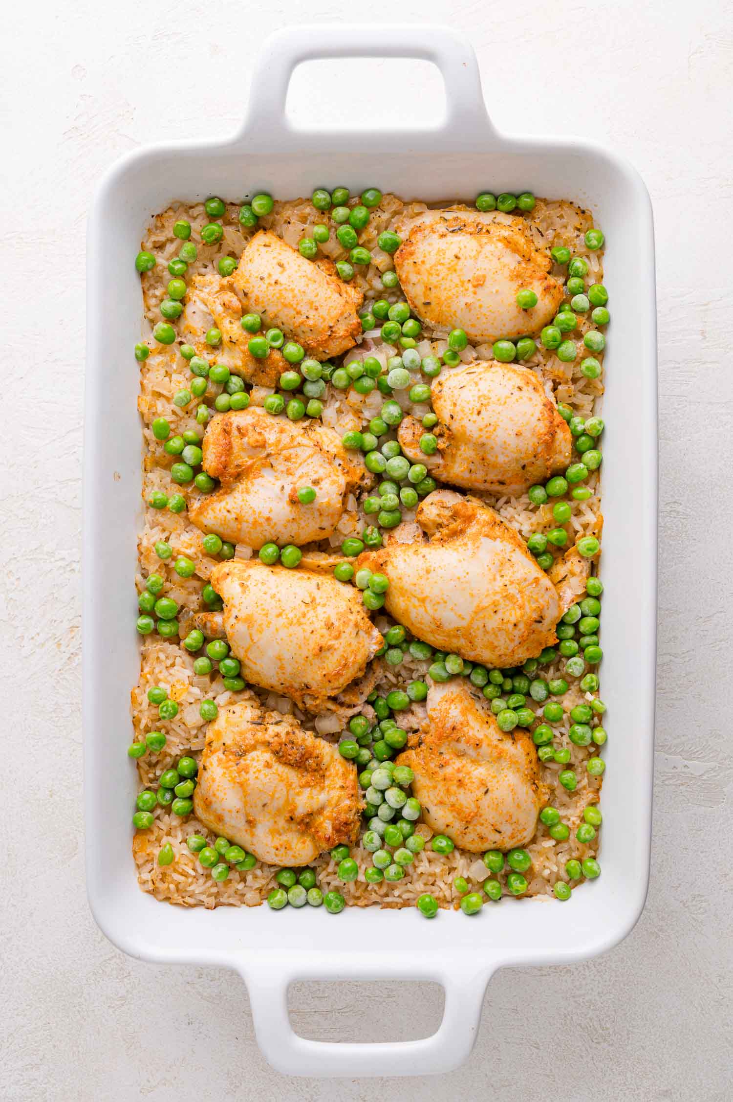 A white baking dish containing baked chicken thighs, green peas, and rice on a light-colored surface.