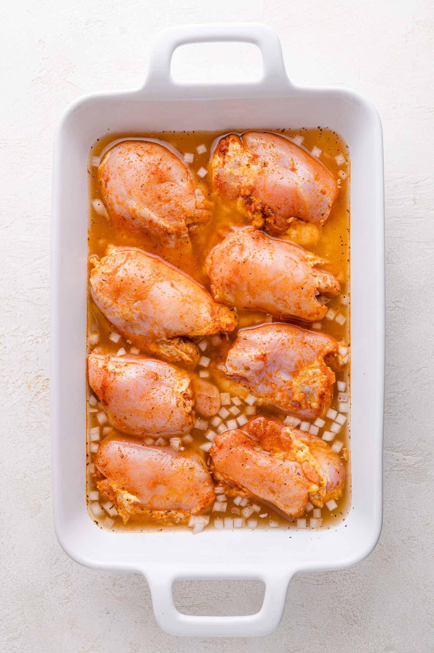 Raw chicken thighs coated with seasoning and placed on chopped onions in a white baking dish with liquid at the bottom, ready to be cooked.