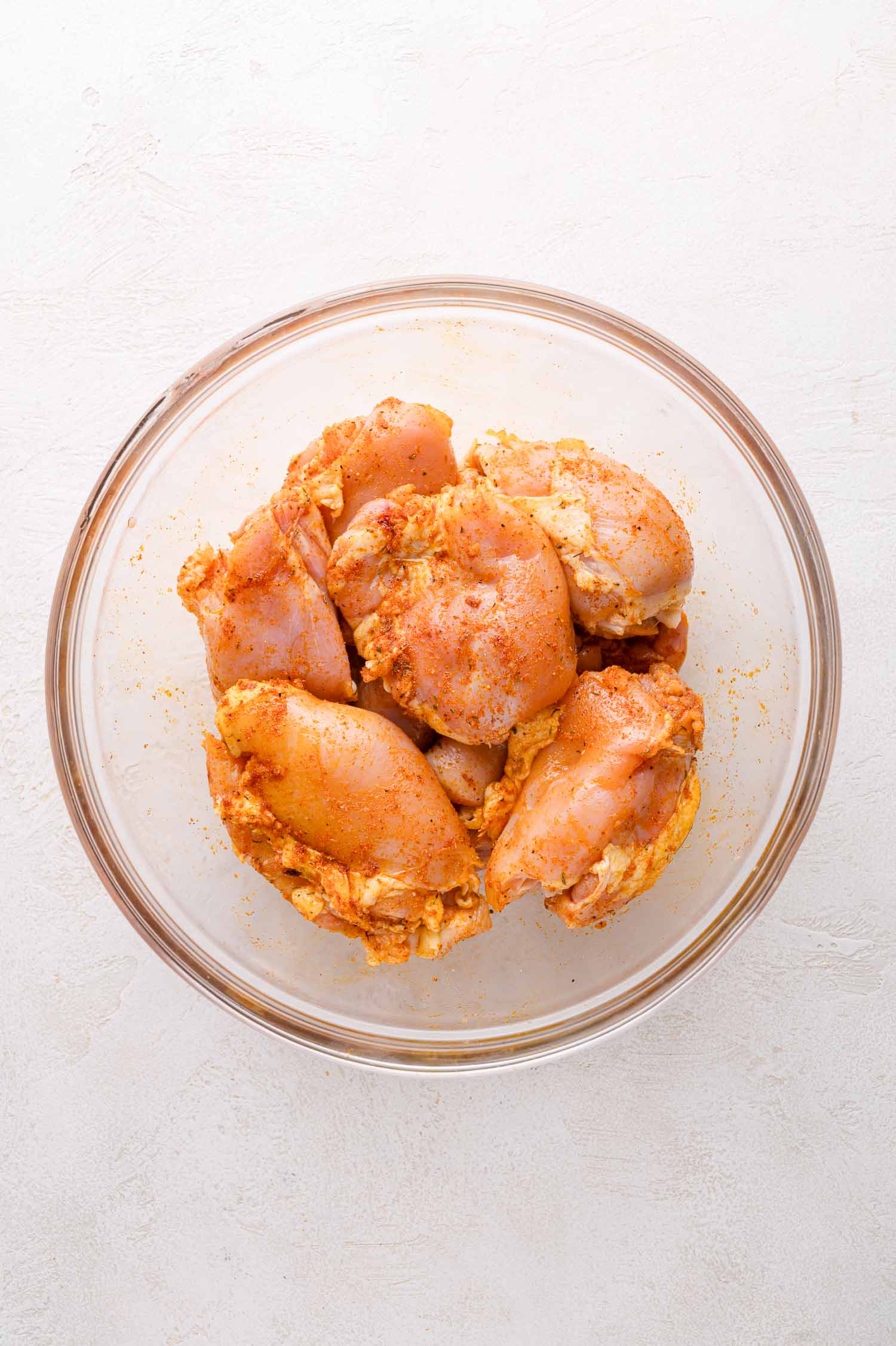 Raw chicken thighs coated in seasoning, placed in a clear glass bowl on a light-colored surface.