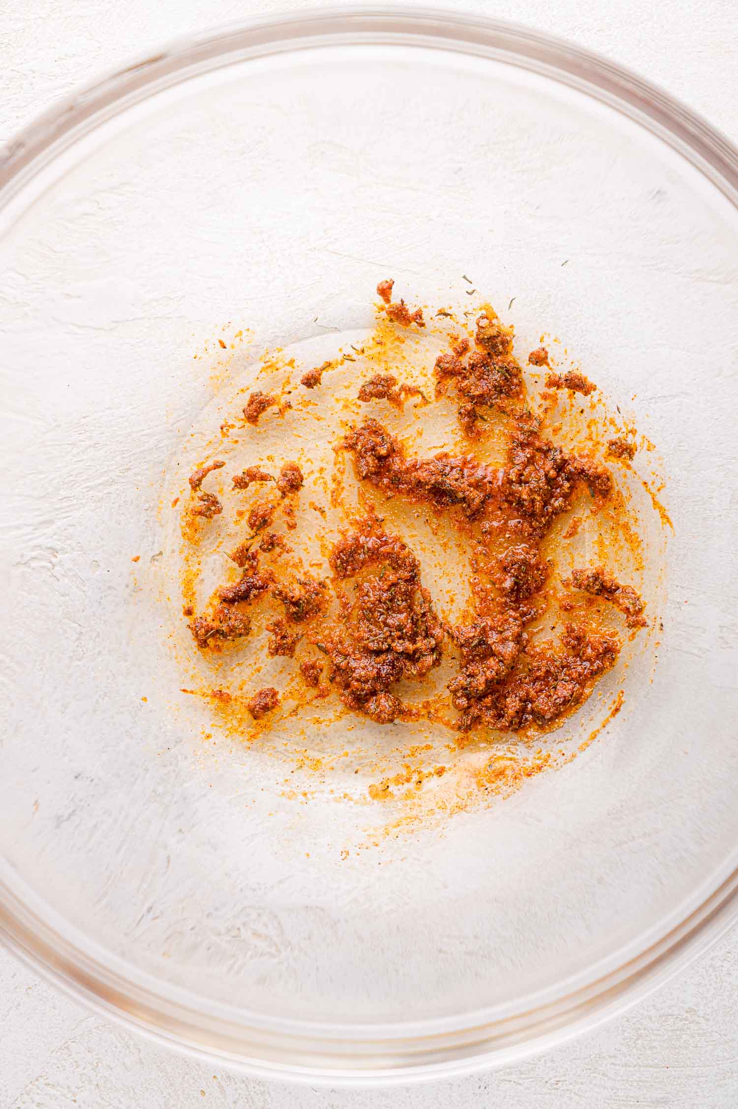 A glass bowl containing a small amount of reddish-brown spice paste, smeared and clumped against the bottom of the bowl.