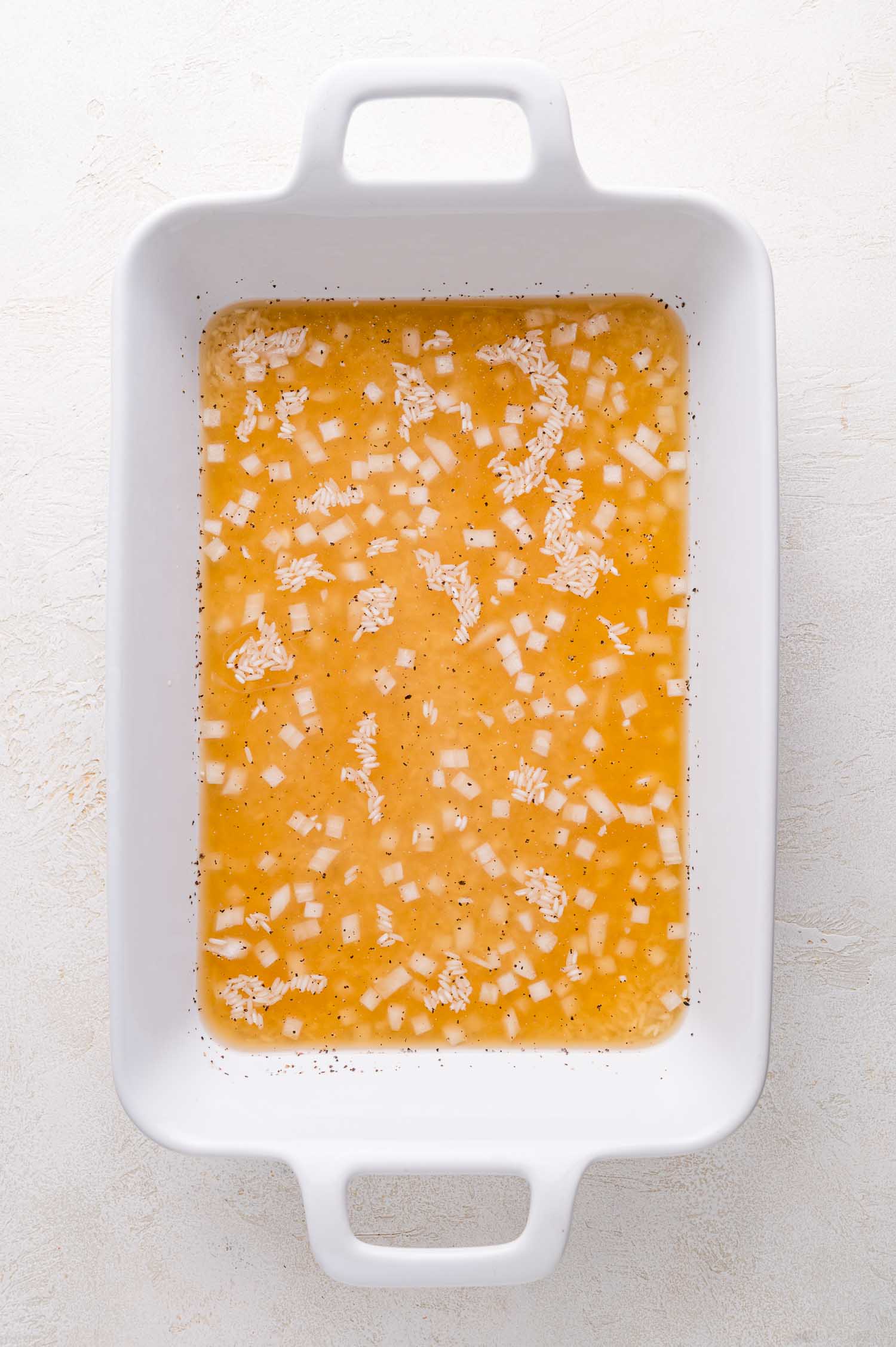 A white rectangular baking dish containing uncooked rice, diced onions, and broth on a light surface.