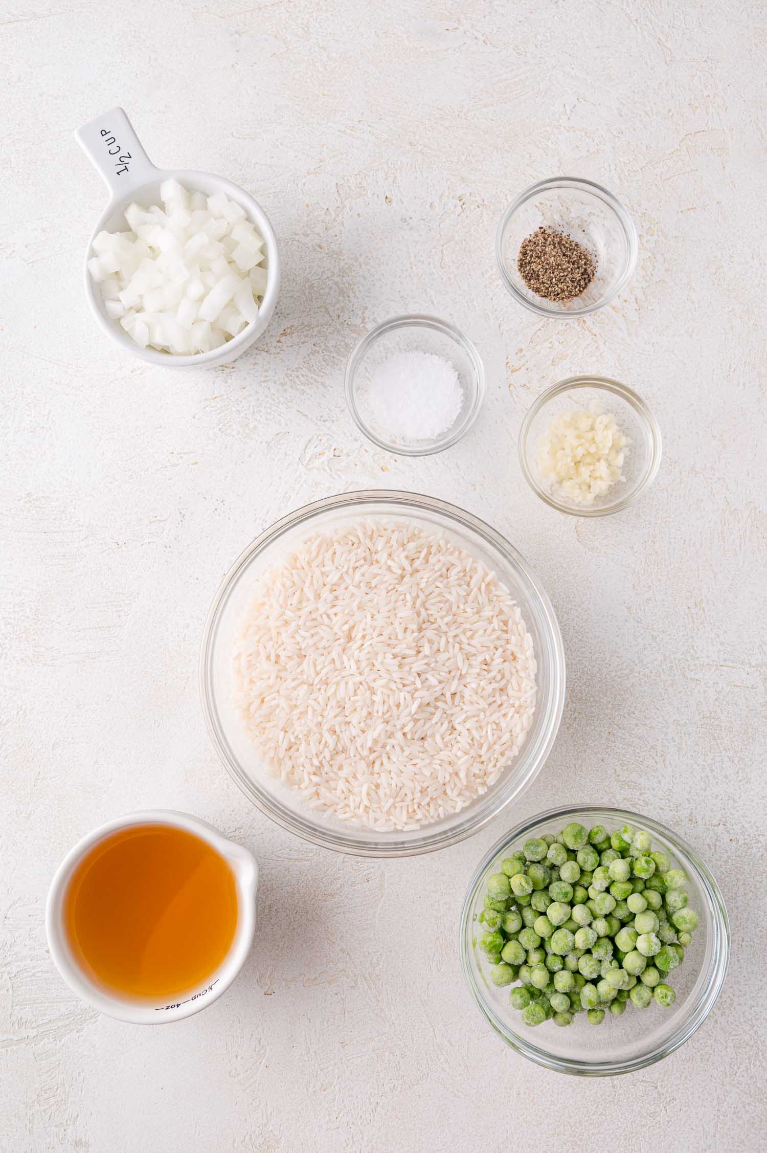 Bowls containing chopped onions, salt, pepper, minced garlic, white rice, frozen peas, and a measuring cup of vegetable broth arranged on a white surface.