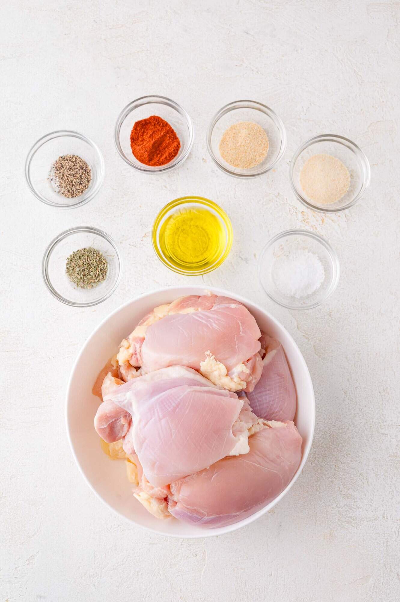 Raw chicken thighs in a white bowl, surrounded by small glass bowls containing oil, salt, black pepper, paprika, garlic powder, onion powder, and dried herbs on a white surface.