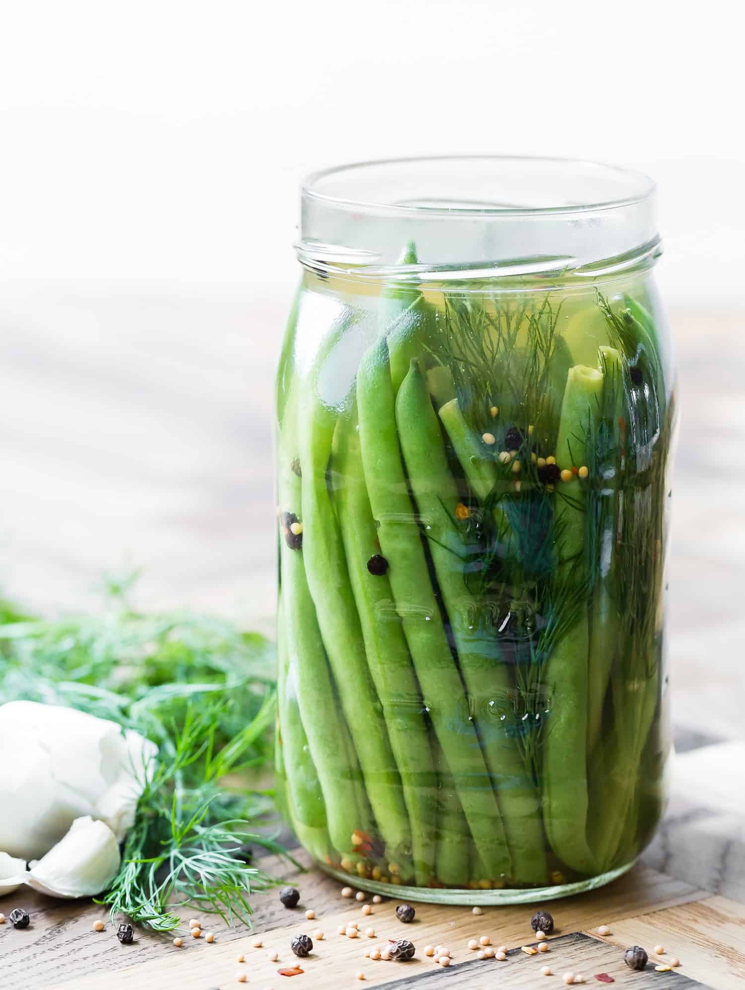 A glass jar filled with pickled green beans, fresh dill, garlic cloves, mustard seeds, and peppercorns, placed on a wooden surface.
