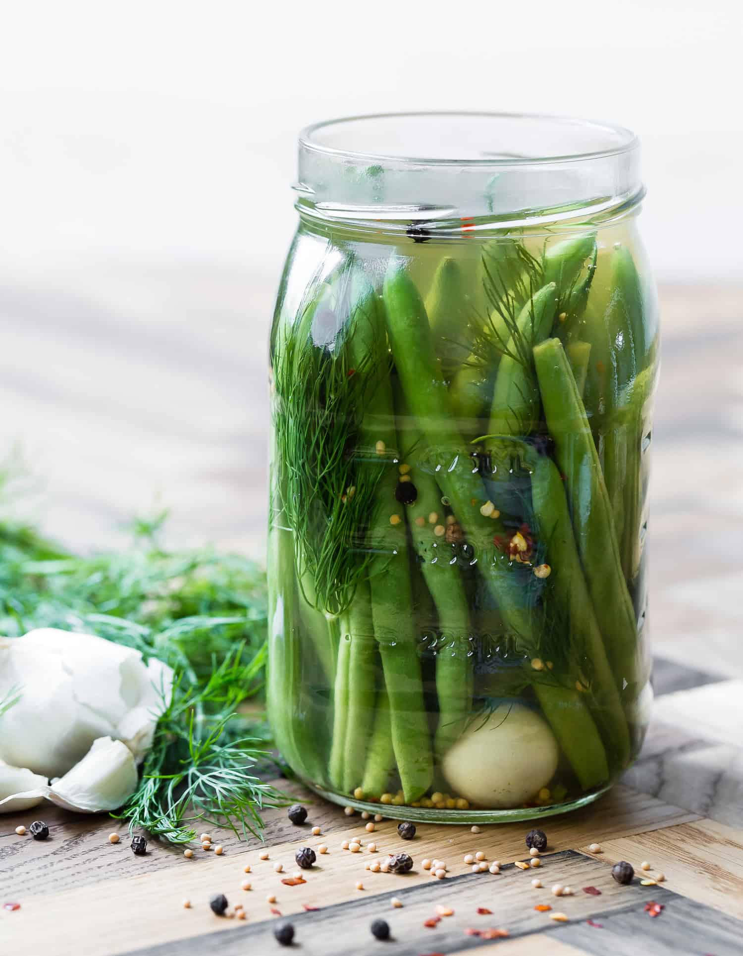 A glass jar filled with pickled green beans, dill, garlic cloves, and various spices, sitting on a wooden surface with fresh dill and garlic nearby.