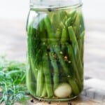 A glass jar filled with pickled green beans, dill, spices, and a garlic clove, sitting on a wooden surface.