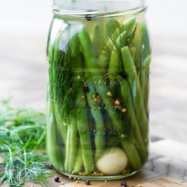 A glass jar filled with pickled green beans, dill, spices, and a garlic clove, sitting on a wooden surface.