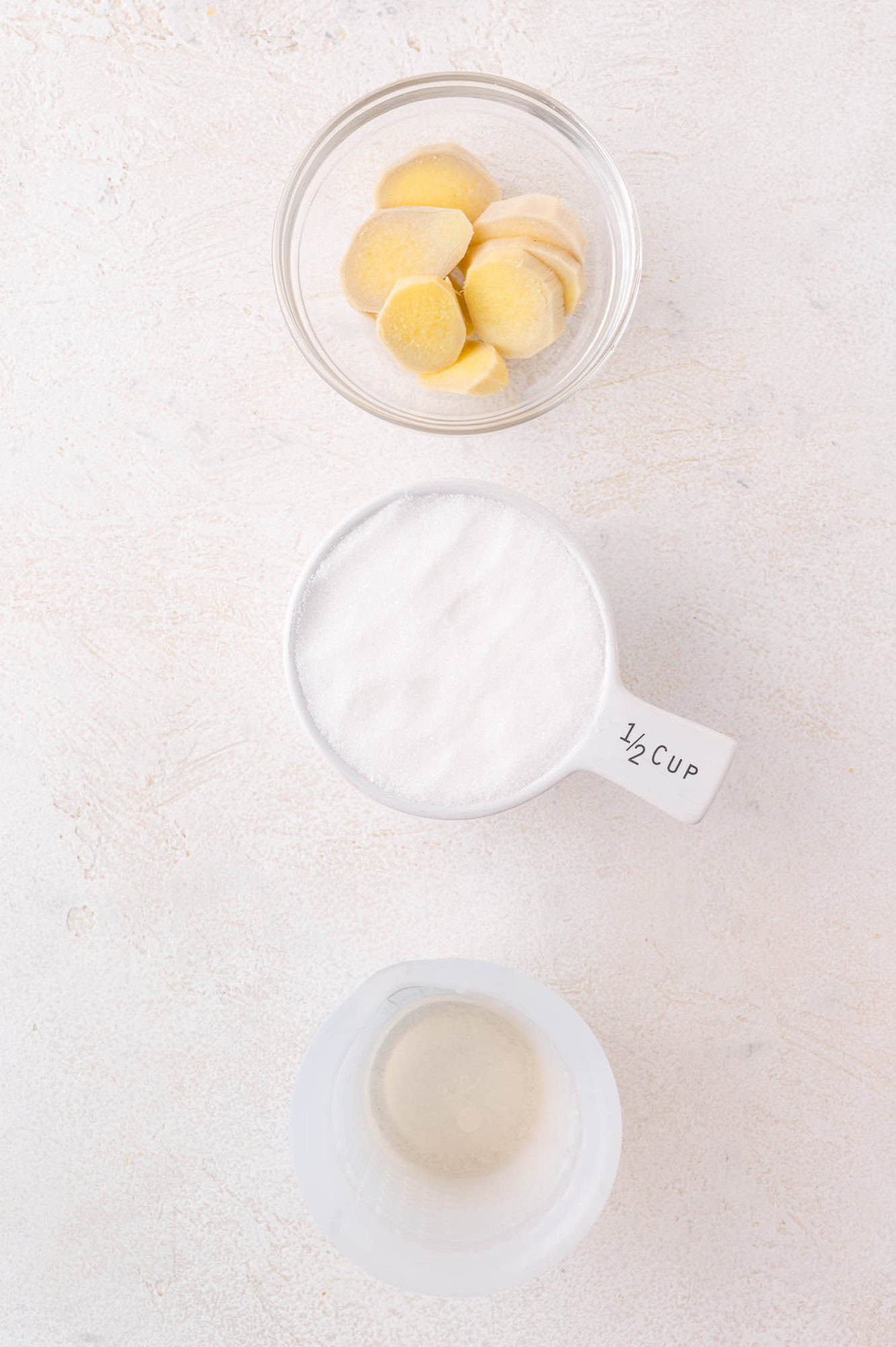 A glass bowl with sliced ginger, a measuring cup with sugar labeled “1/2 cup,” and a small jug of clear liquid arranged on a light surface.