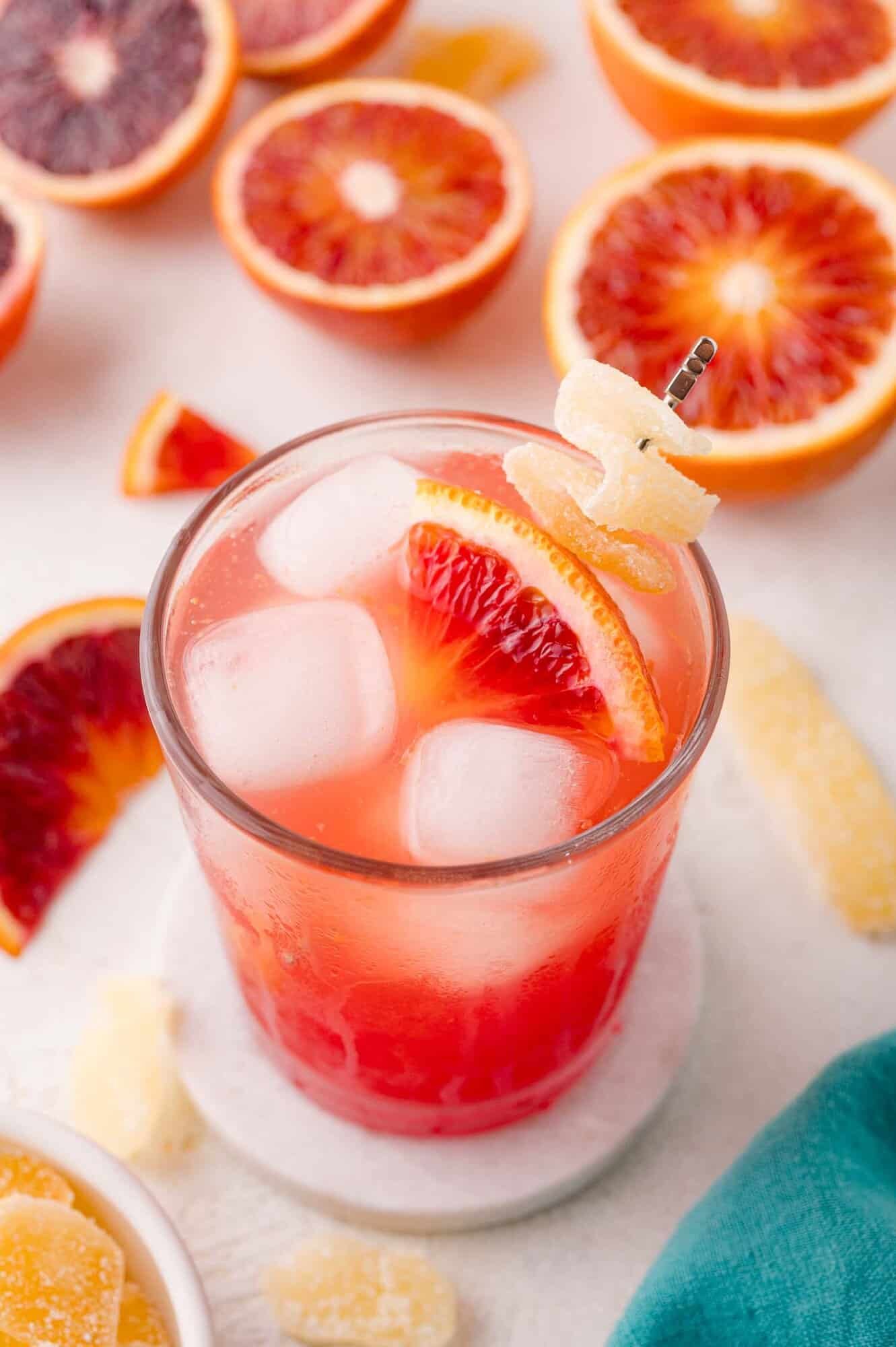 A glass of iced blood orange mocktail garnished with a blood orange slice and candied ginger, surrounded by sliced blood oranges and ginger pieces.