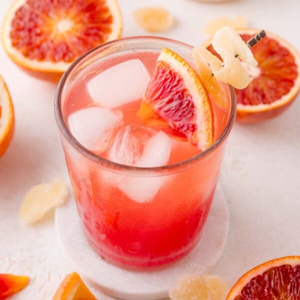 A glass of blood orange mocktail with ice, garnished with a blood orange wedge and candied ginger, sits on a white coaster surrounded by sliced blood oranges and ginger pieces.