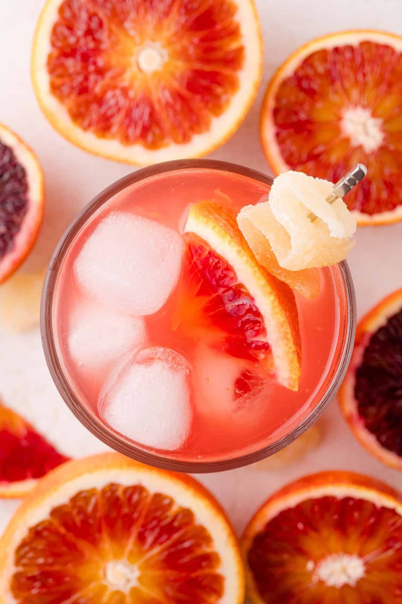 A glass of blood orange mocktail with ice cubes, garnished with a slice of blood orange and a twist of candied ginger, surrounded by halved blood oranges.
